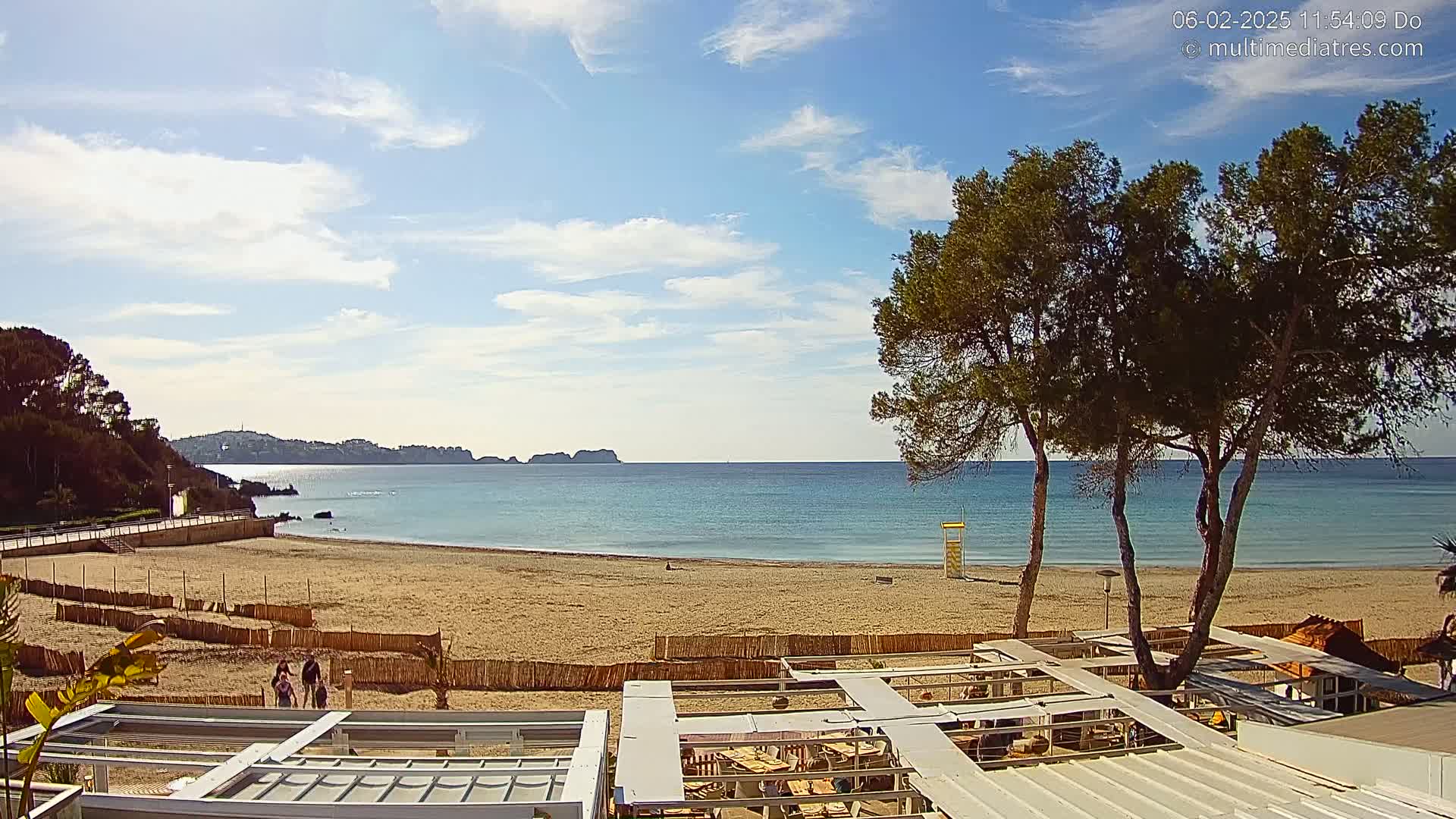 A sandy beach with calm, light blue water stretches to a distant coastline under a partly cloudy sky, with several pine trees and outdoor seating visible in the foreground.