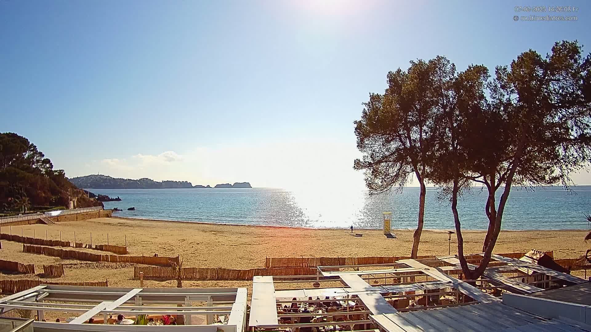 A sandy beach with calm, clear blue water under a sunny sky is partially enclosed by a wooden fence, with several pine trees and an outdoor restaurant visible in the foreground.