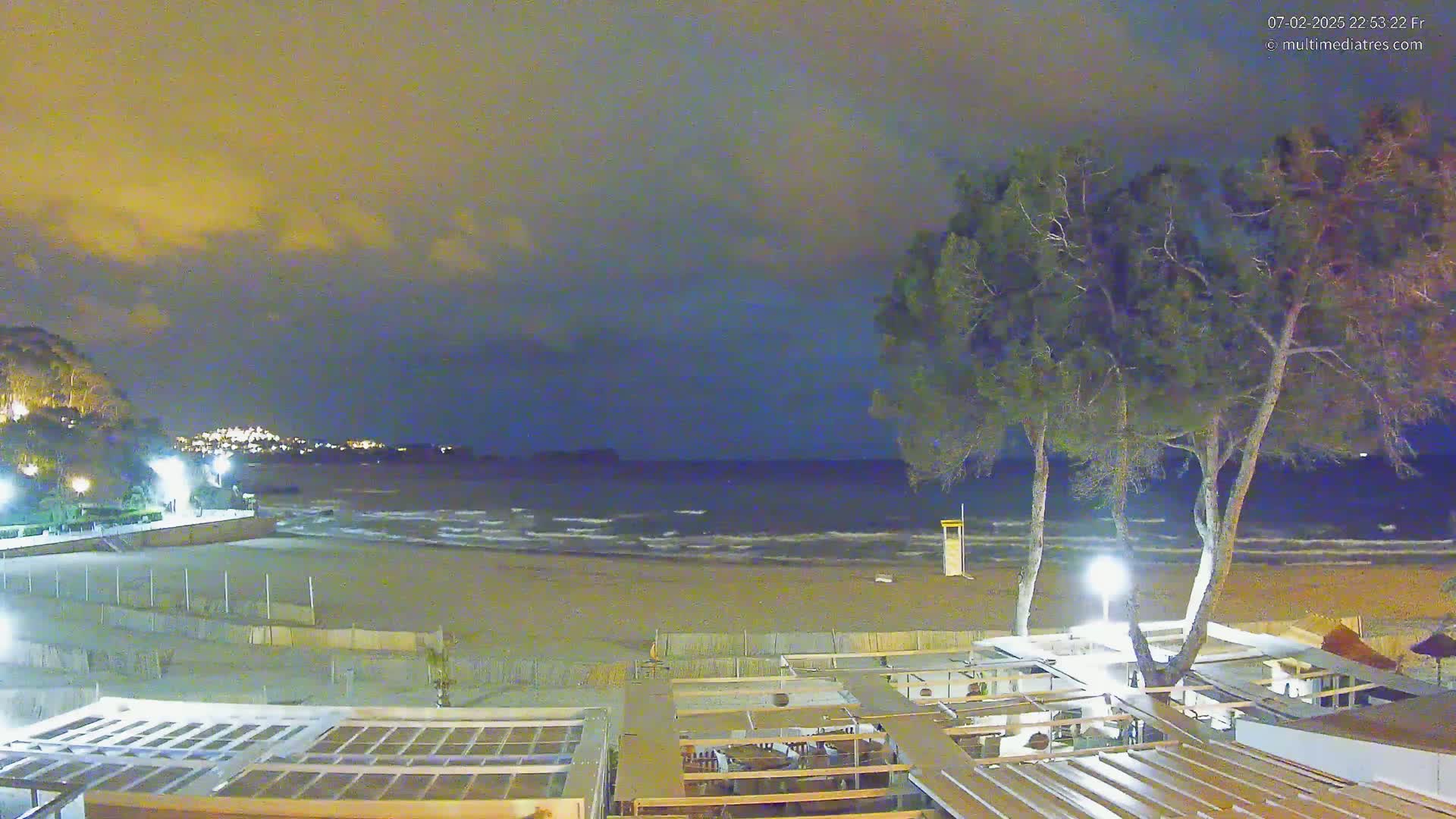 A nighttime view of a beach with gentle waves under a cloudy sky, showing a sandy shore, beachfront buildings, and tall trees.