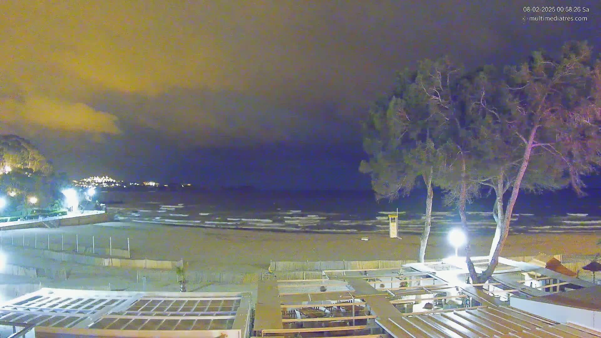 A nighttime view of a beach with gently lapping waves under a cloudy, dark-yellow sky, showing a beachfront building and several tall trees.