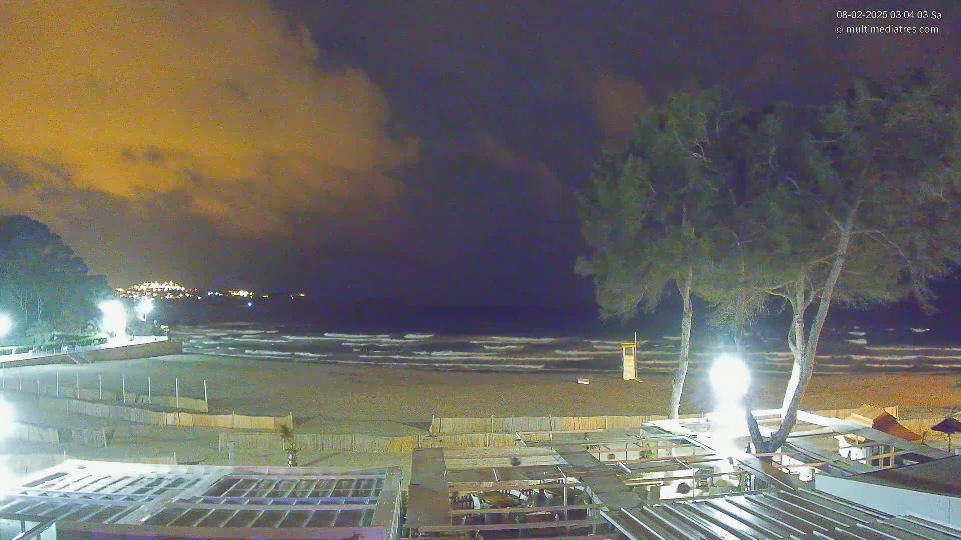 A nighttime beach scene with gently rolling waves under a cloudy, orange-hued sky shows a sandy beach with buildings and palm trees.