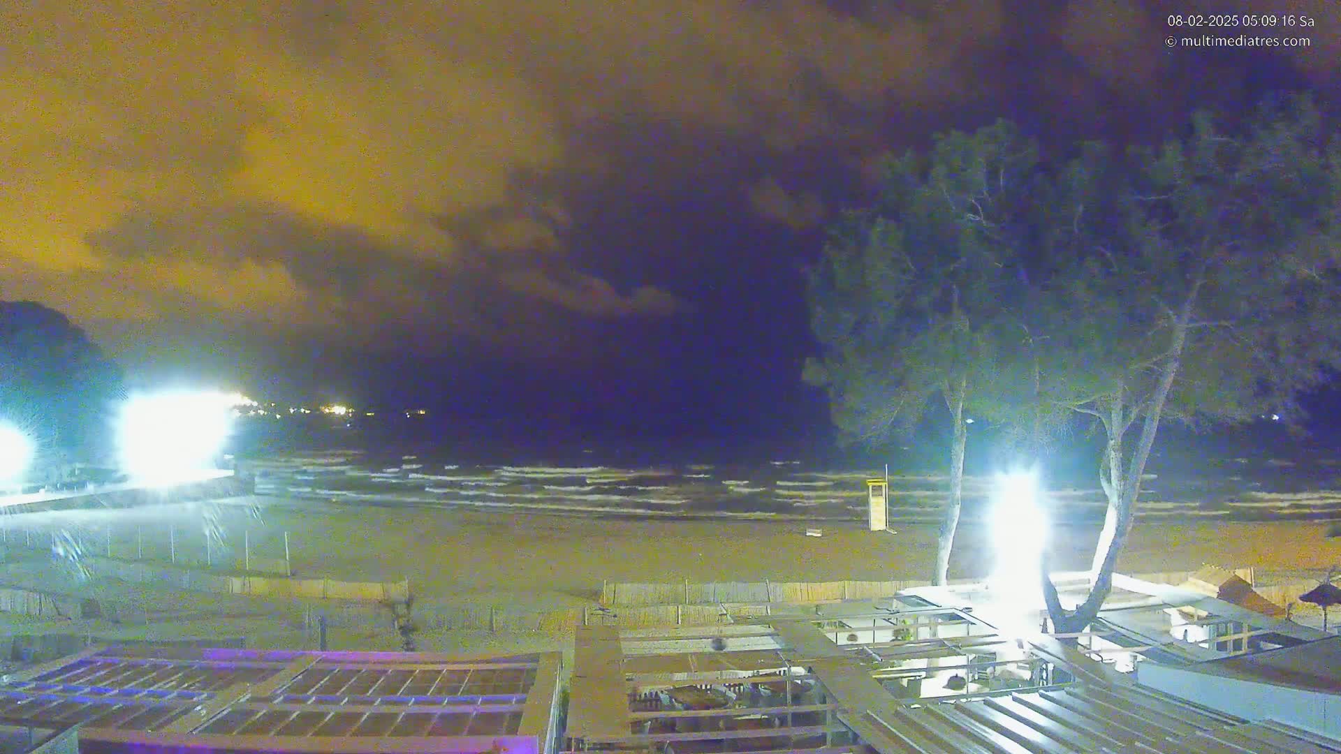 A nighttime beach scene with moderate waves under a cloudy, dark yellow-orange sky shows a sandy beach, a shoreline building with lights, and two tall trees in the foreground.