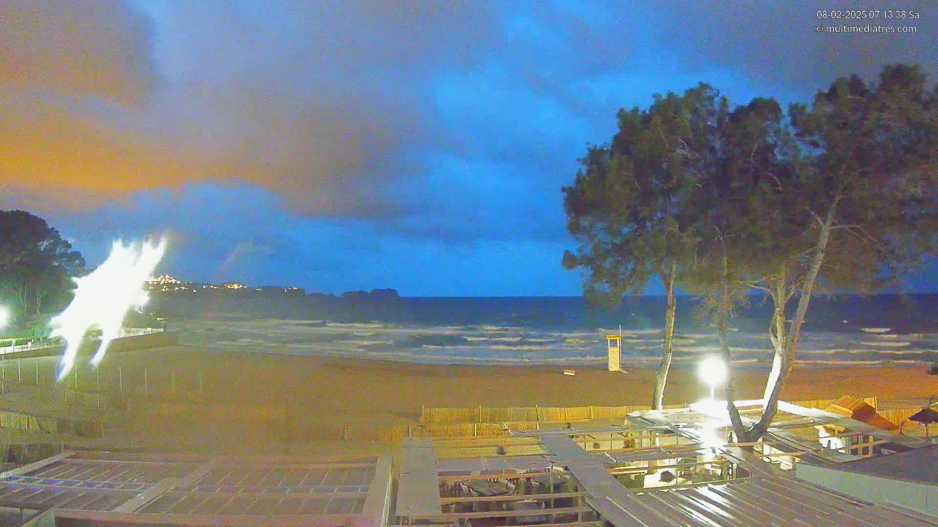 A sandy beach with a choppy sea under a twilight sky is viewed from a rooftop patio, partially obscured by several pine trees.