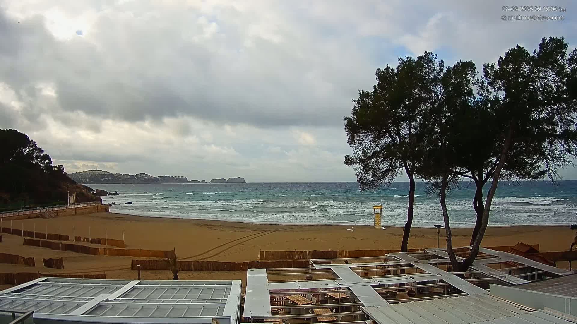 A sandy beach with moderate waves under a cloudy sky is viewed from above a partially covered outdoor patio area, with dark green pine trees in the foreground.