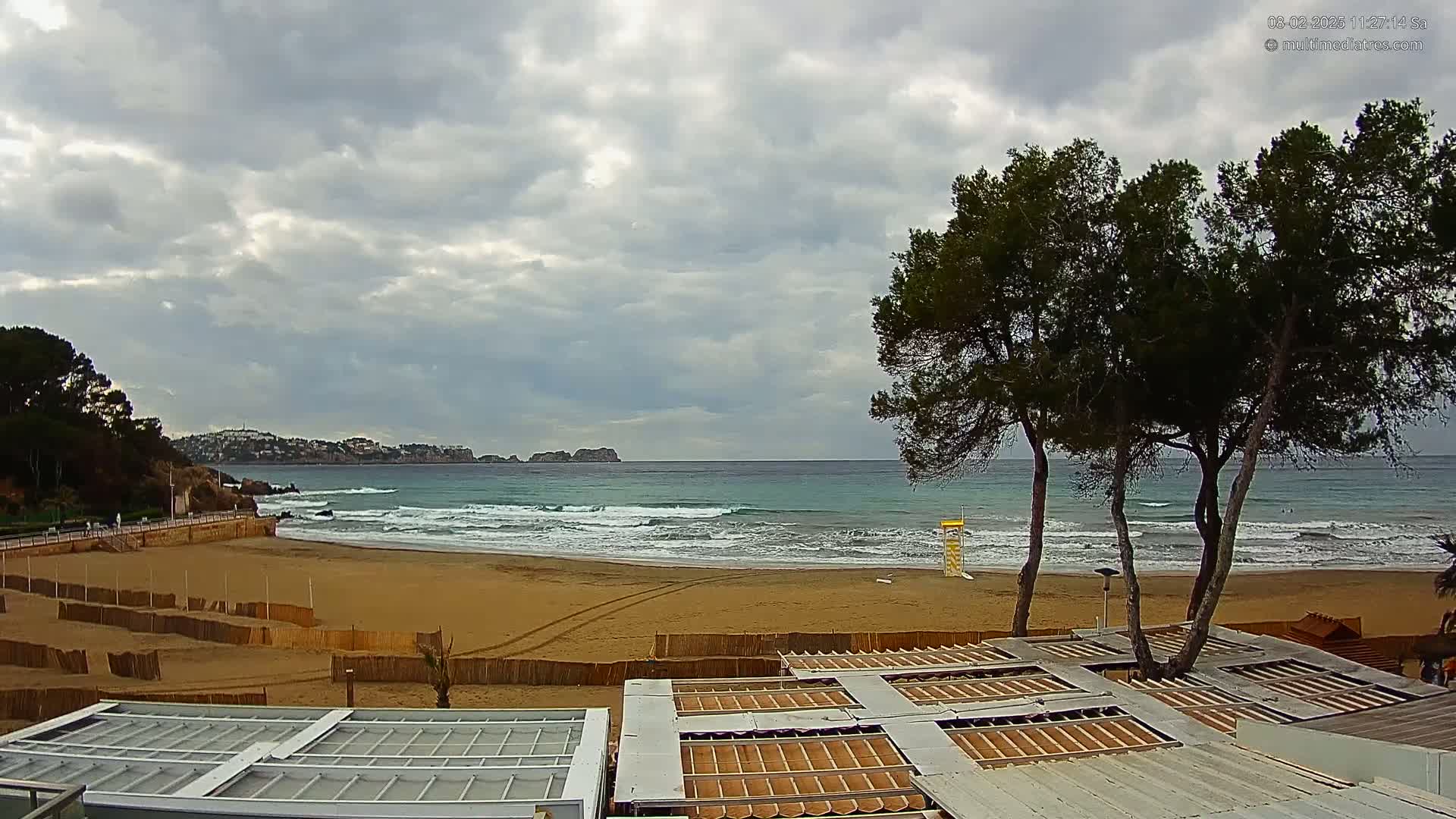 A sandy beach with moderate waves under a cloudy sky is bordered by green trees and beach structures.