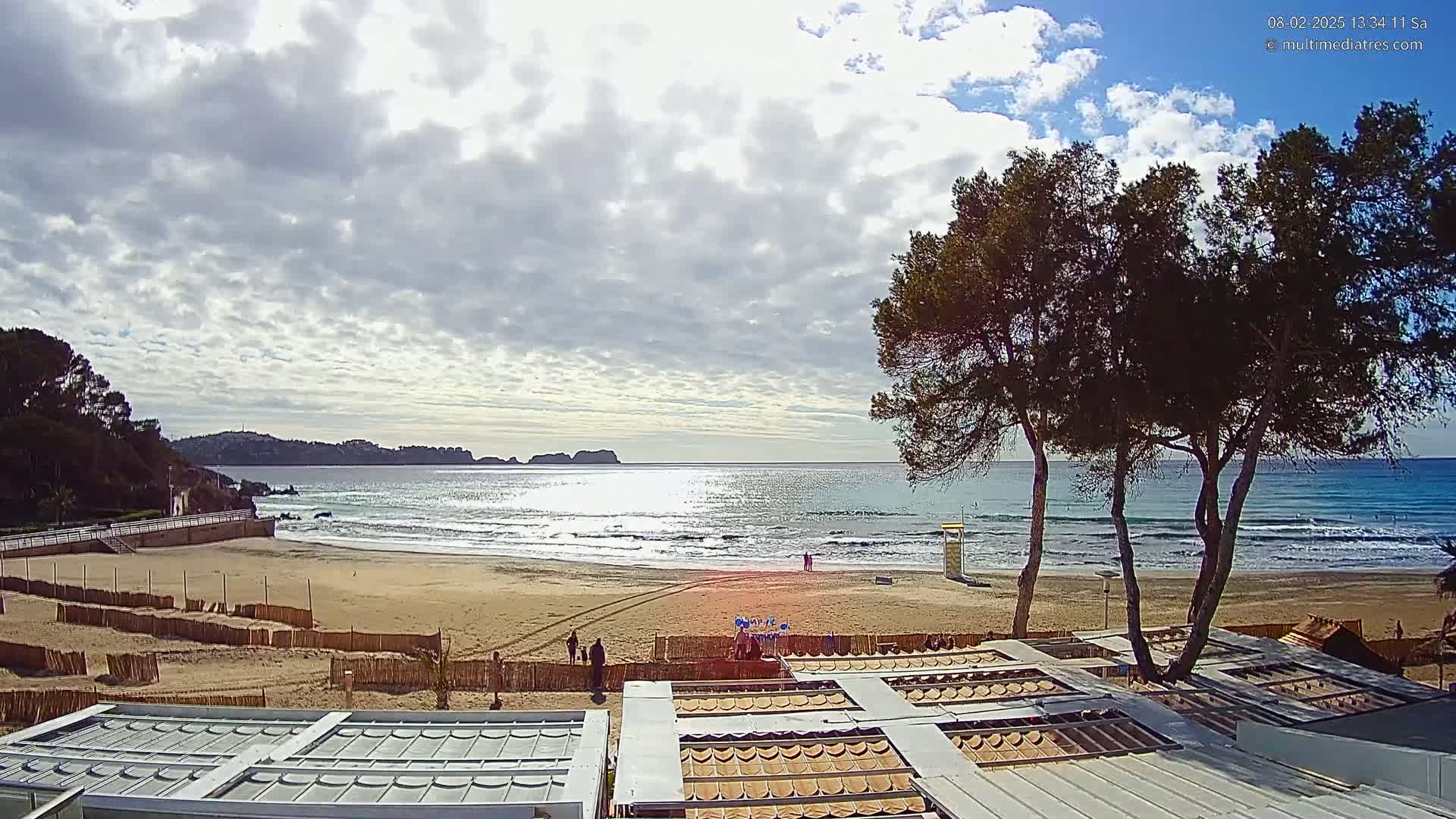 A sandy beach with calm waves under a partly cloudy sky is viewed from above, with several trees in the foreground and buildings partially visible.