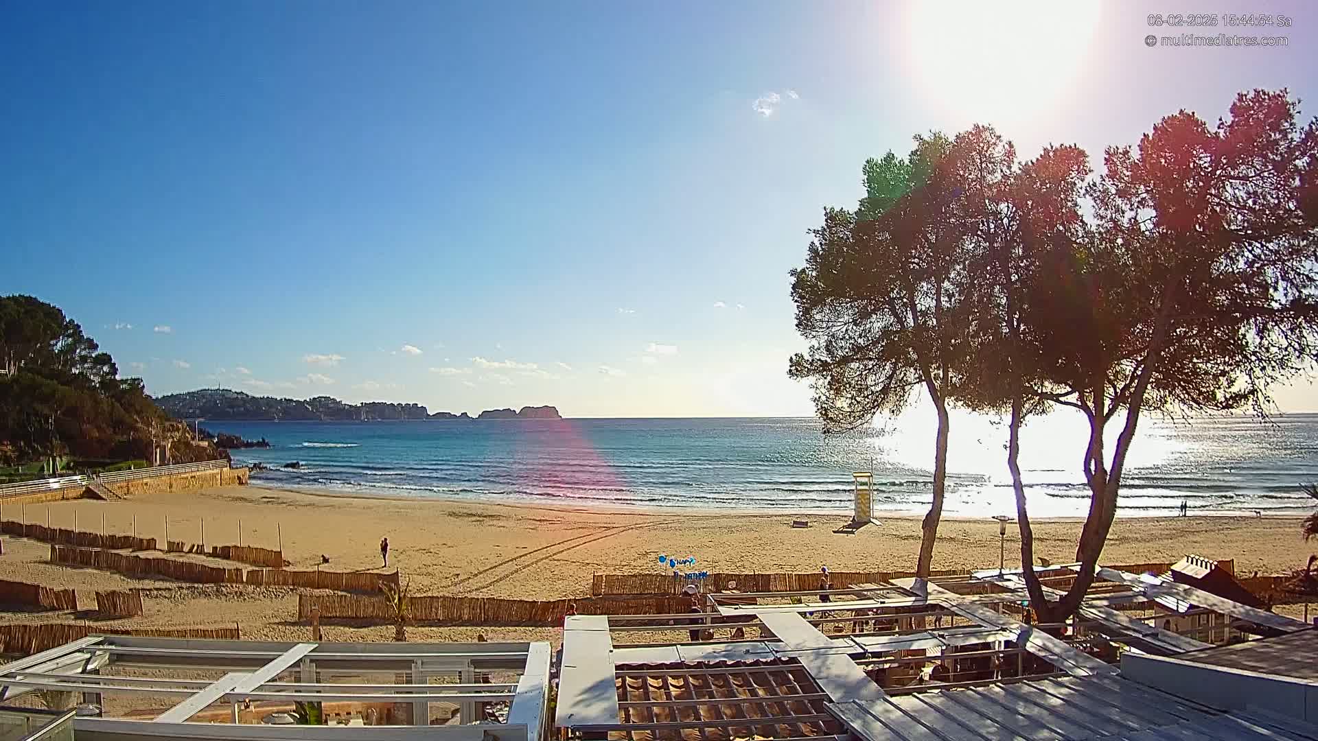 A sandy beach with calm ocean waves under a sunny sky is viewed from above a partially shaded outdoor patio area, with several trees in the foreground.