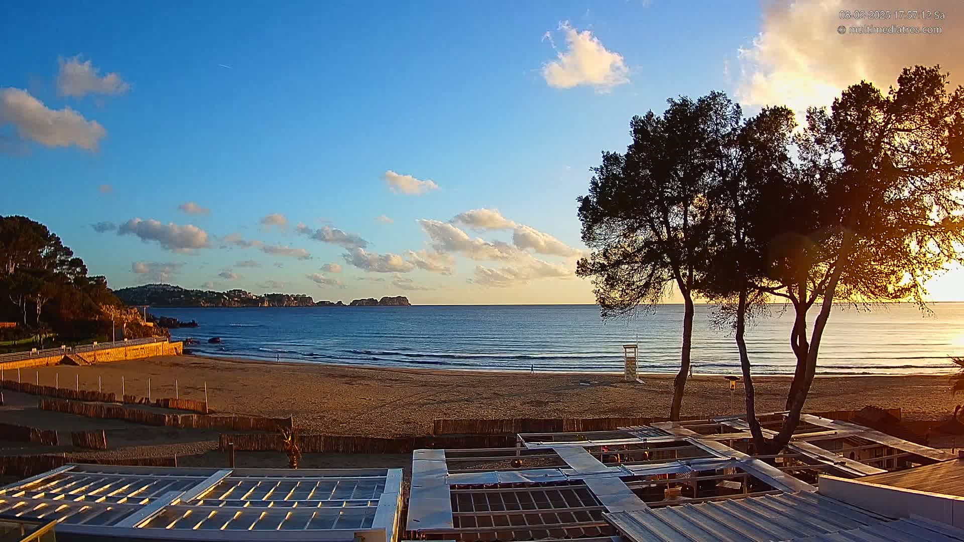 A sandy beach with gentle waves and a calm sea at sunset, partially obscured by several pine trees and beachside structures.