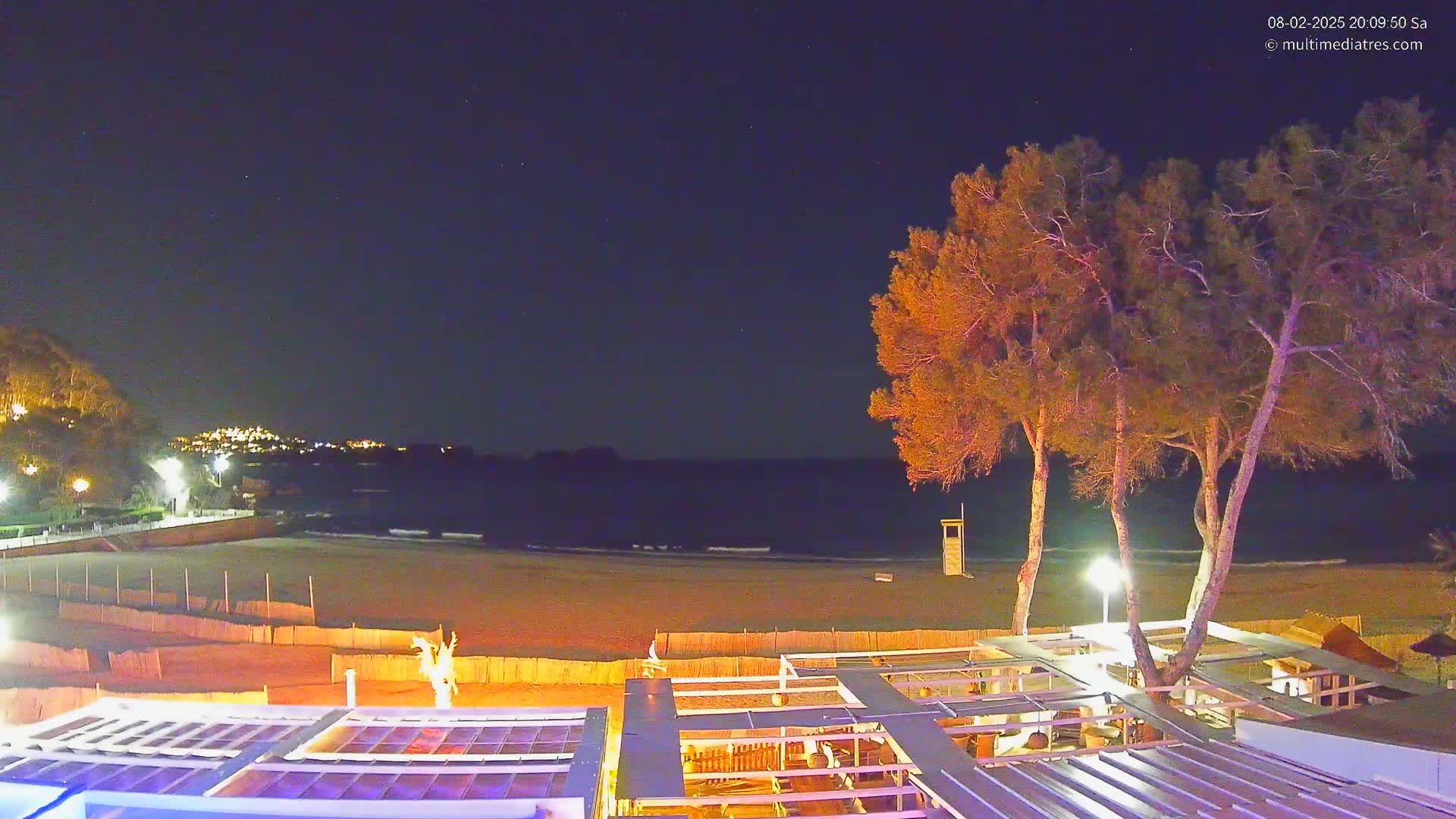 A nighttime view of a sandy beach, calm ocean, and a beachfront establishment under a clear, starry sky.