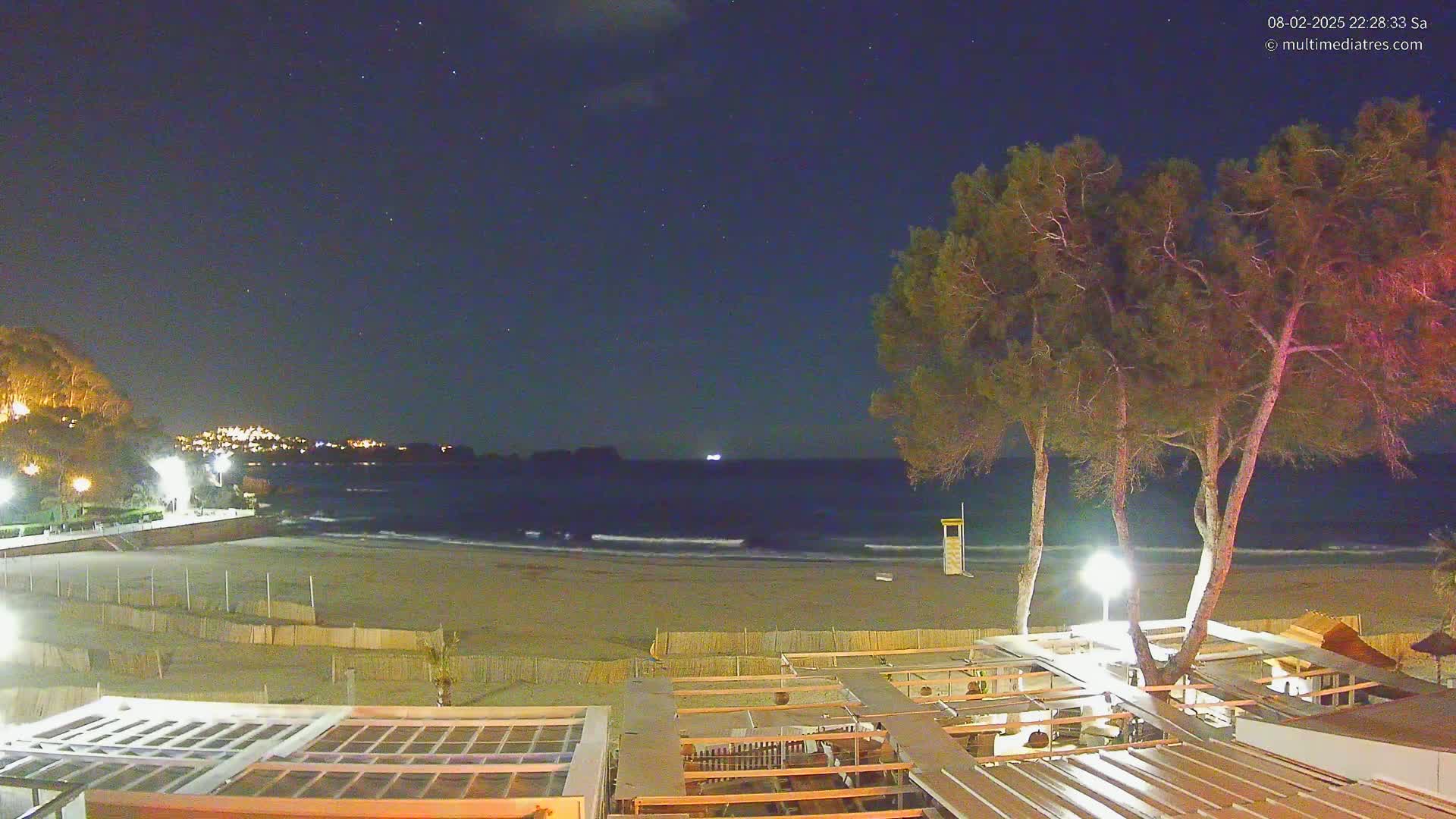 A nighttime view of a sandy beach with calm waves under a clear, starry sky, showing a beachfront structure and trees in the foreground.