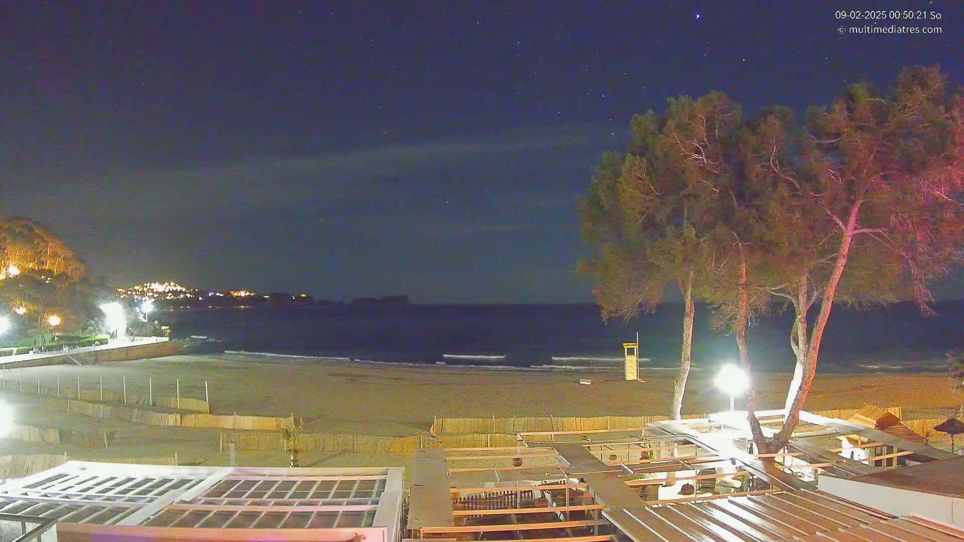 A nighttime beach scene shows a sandy shore, calm ocean waves, a lifeguard stand, several trees, and a building with a partially covered patio under a clear, starry sky.