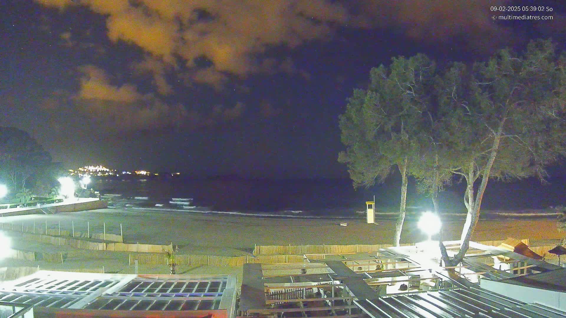 A nighttime view of a beach with a dark, cloudy sky, gently lapping waves, and several illuminated trees near a beachfront building.
