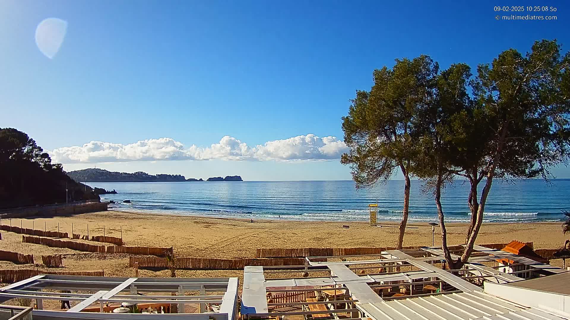 A sandy beach with gentle waves under a mostly sunny sky, viewed from a patio overlooking the ocean and several trees.