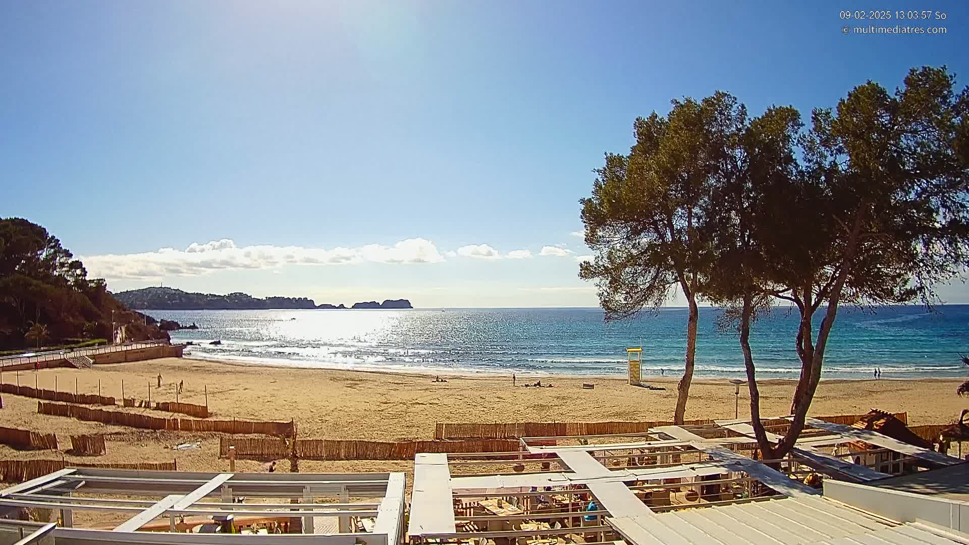 A sandy beach with calm waves under a mostly sunny sky is partially enclosed by a wooden fence; a few people are visible on the beach, and there are several trees in the foreground, with the tops of some buildings in the lower portion of the image.