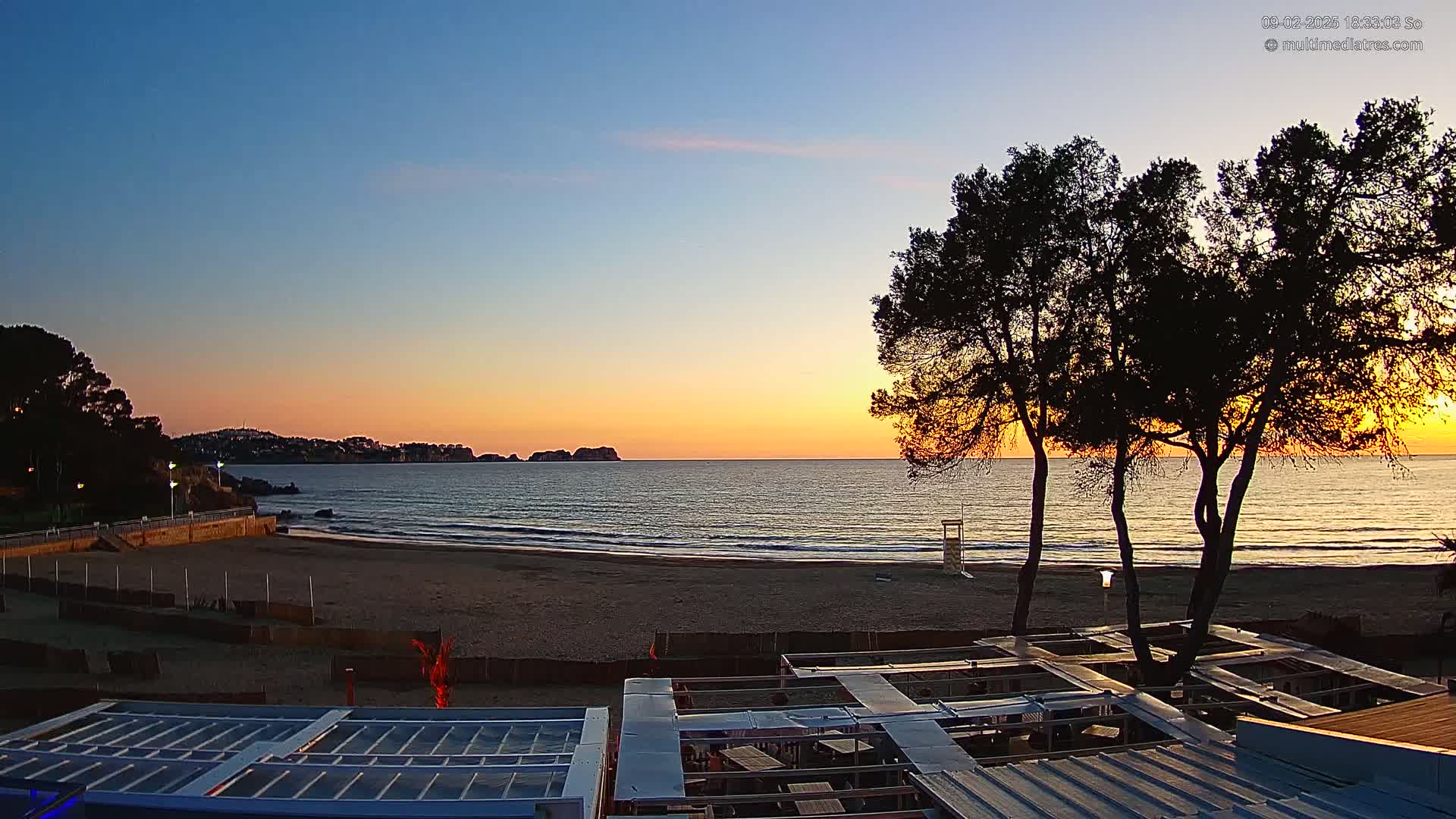 A beach at sunset, with calm ocean waves lapping the shore, is partially obscured by several dark trees in the foreground and the rooftops of beachfront structures.