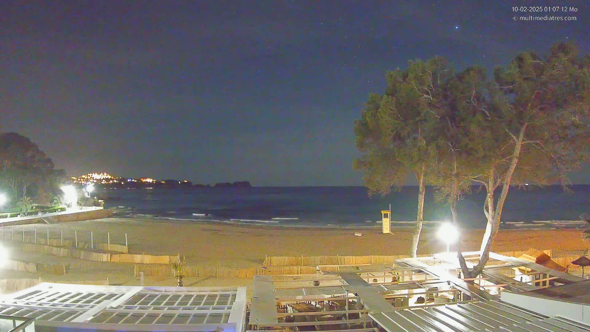 A nighttime view of a sandy beach with calm waves under a clear, starry sky, featuring a beachfront restaurant and several trees.