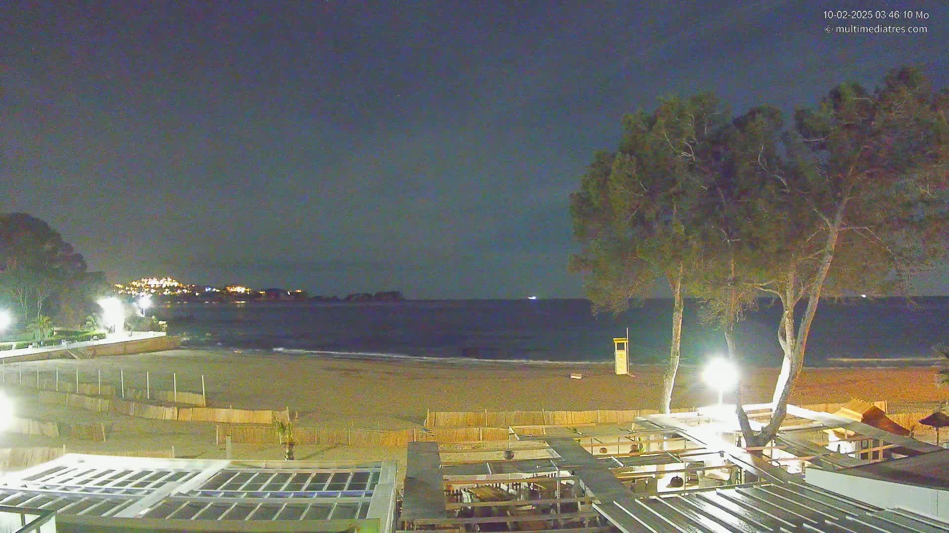 A nighttime view of a sandy beach with gentle waves, a lifeguard stand, and a restaurant structure under a clear, dark sky.