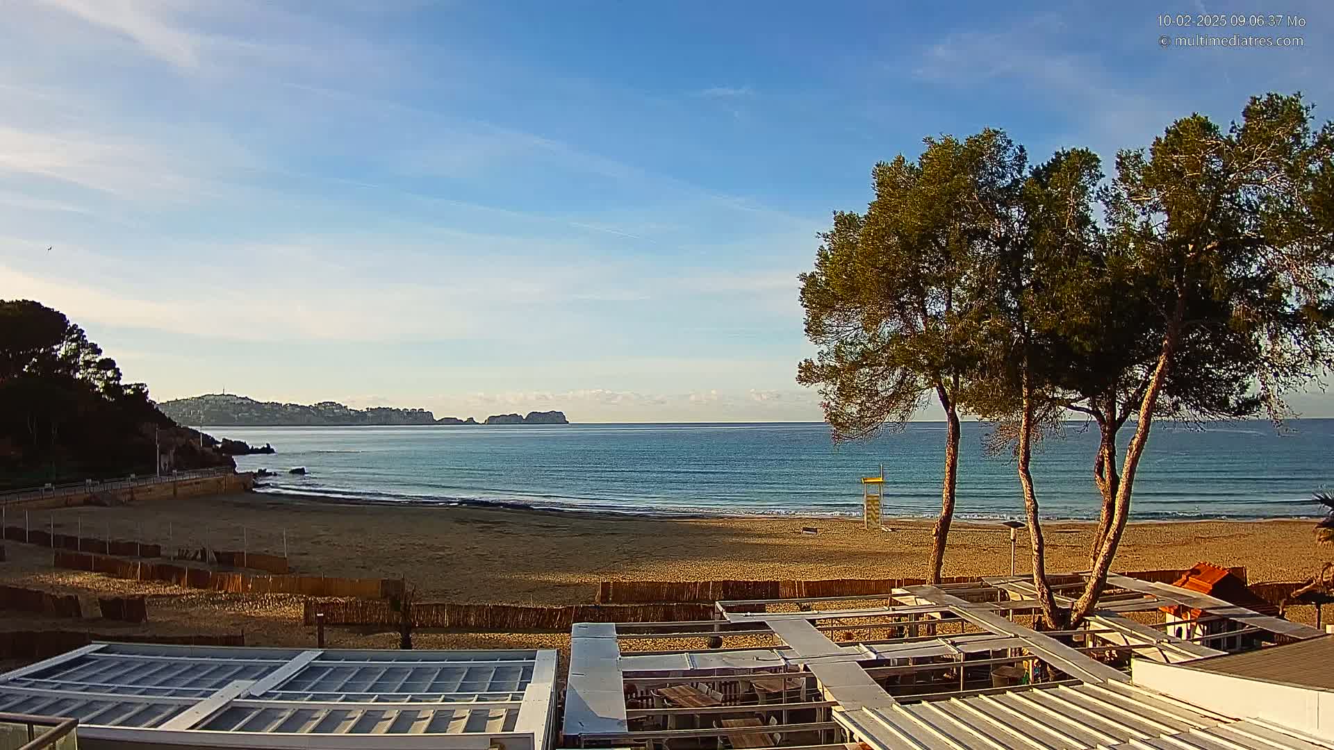 A sandy beach with calm ocean waves stretches out to a distant coastline under a mostly clear sky, with several pine trees in the foreground partially obscuring a beachside structure.