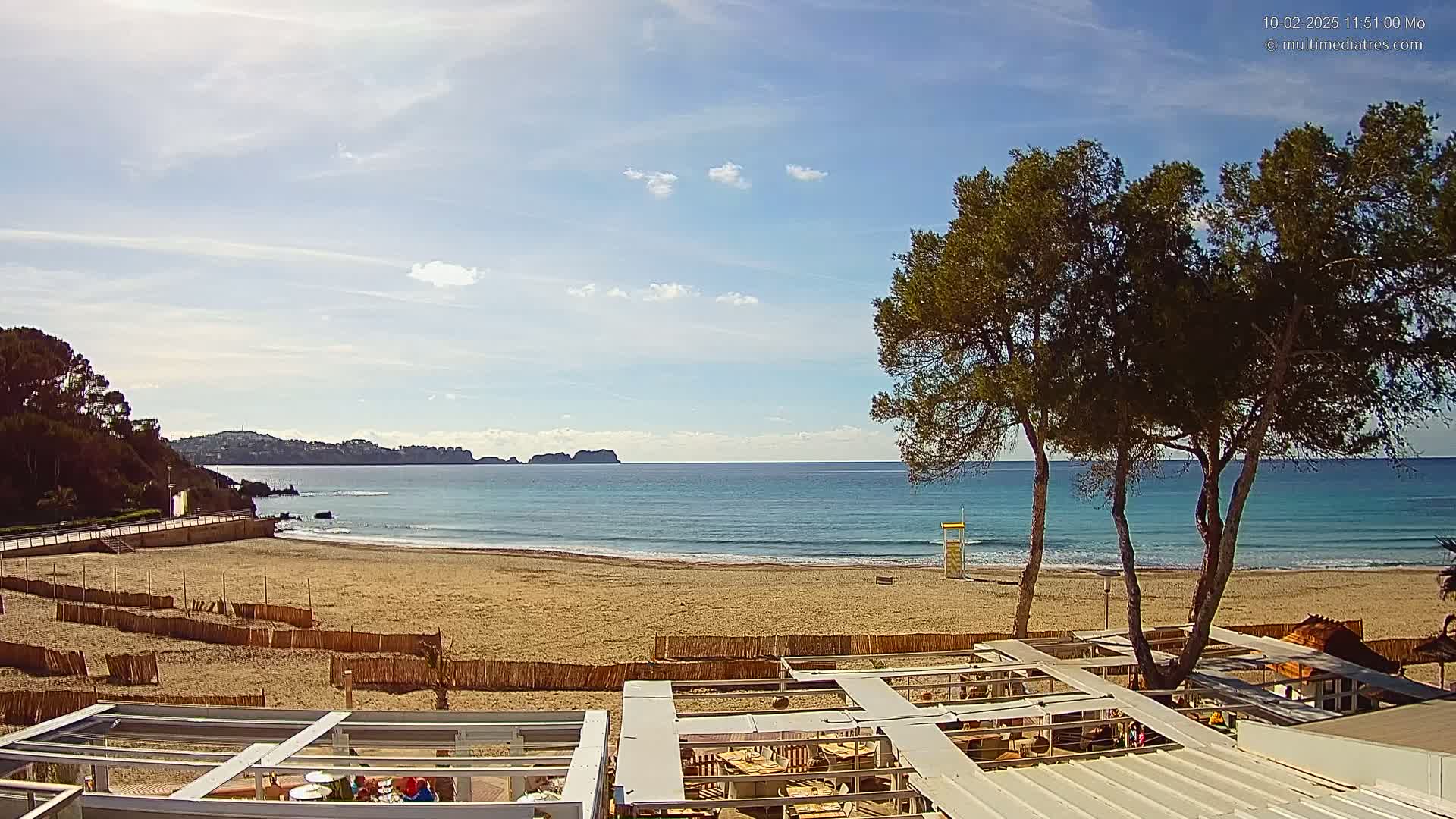 A sandy beach with calm ocean waves under a mostly sunny sky is viewed from an elevated position overlooking outdoor patio furniture.