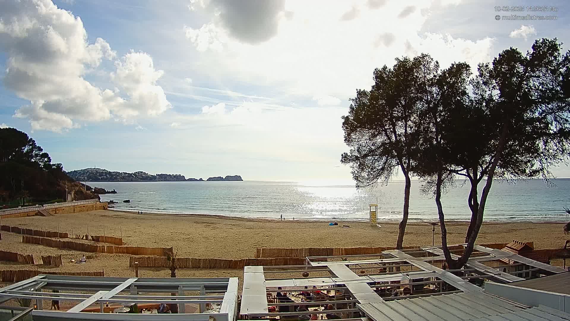 A sandy beach with calm ocean water under a partly cloudy sky is viewed from an elevated position, showing a beachfront restaurant with patrons and several pine trees.