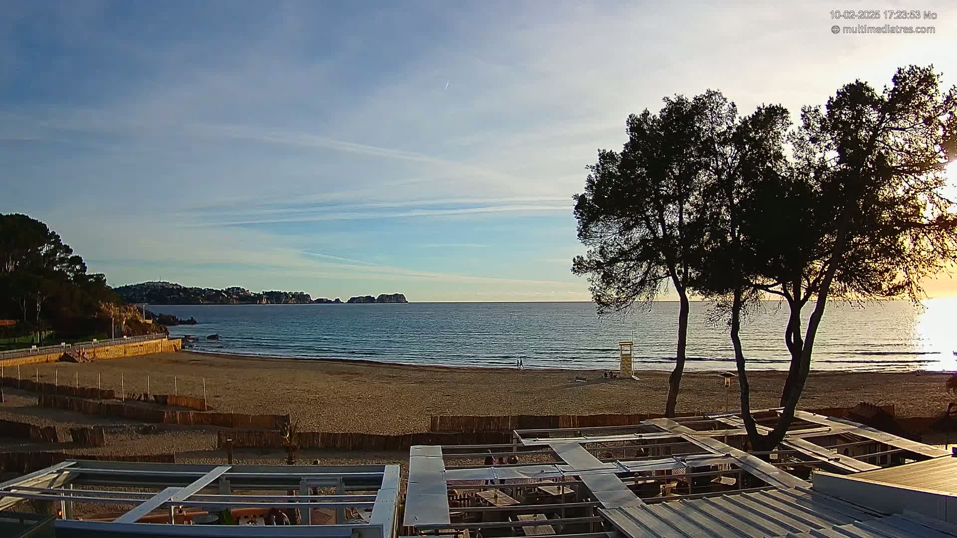 A sandy beach with calm ocean water and a clear sky at sunset, partially obscured by several silhouetted pine trees and the tops of outdoor structures.