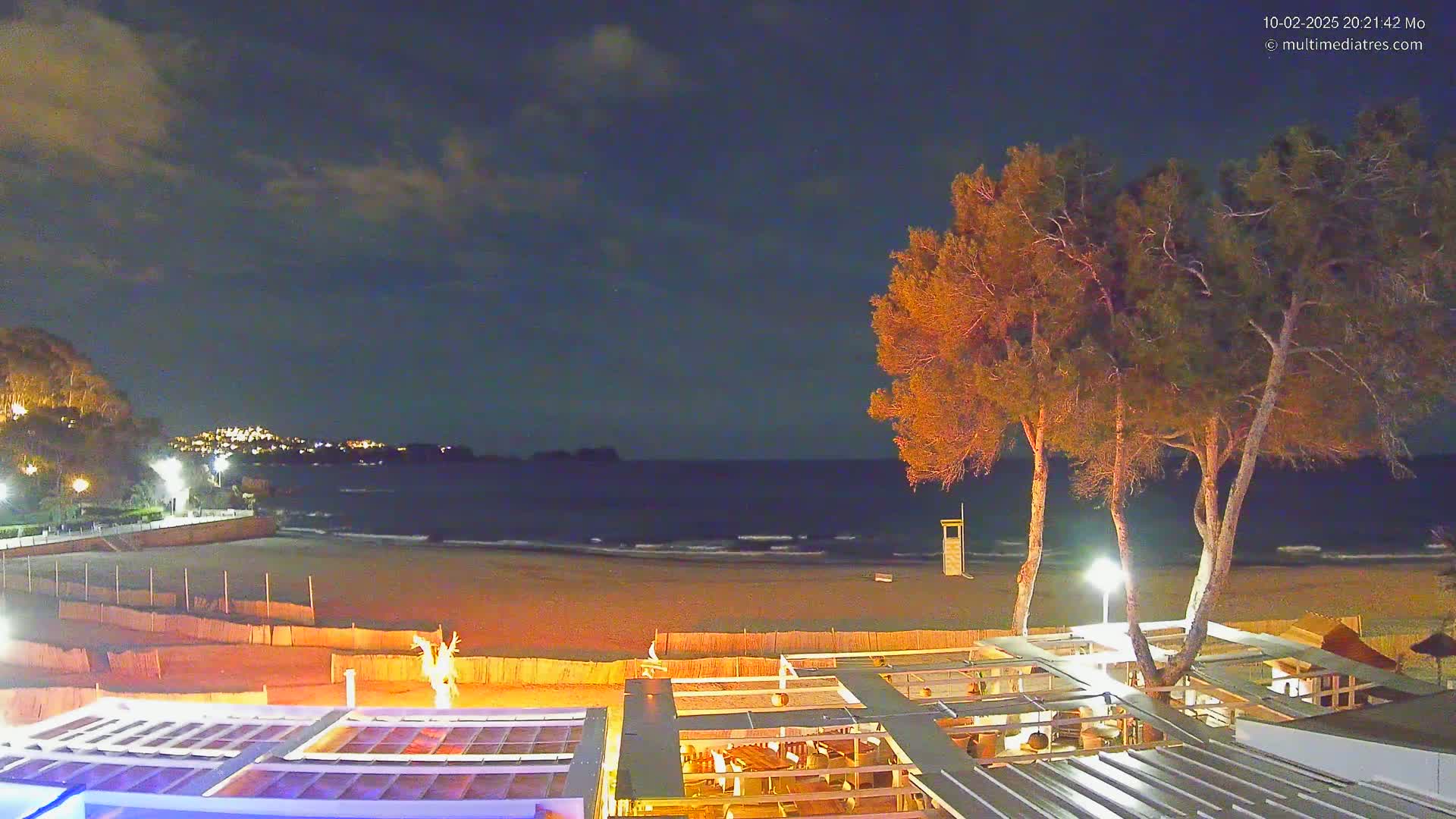 A nighttime view of a sandy beach with gentle waves, illuminated buildings, and several tall, illuminated trees under a partly cloudy sky.
