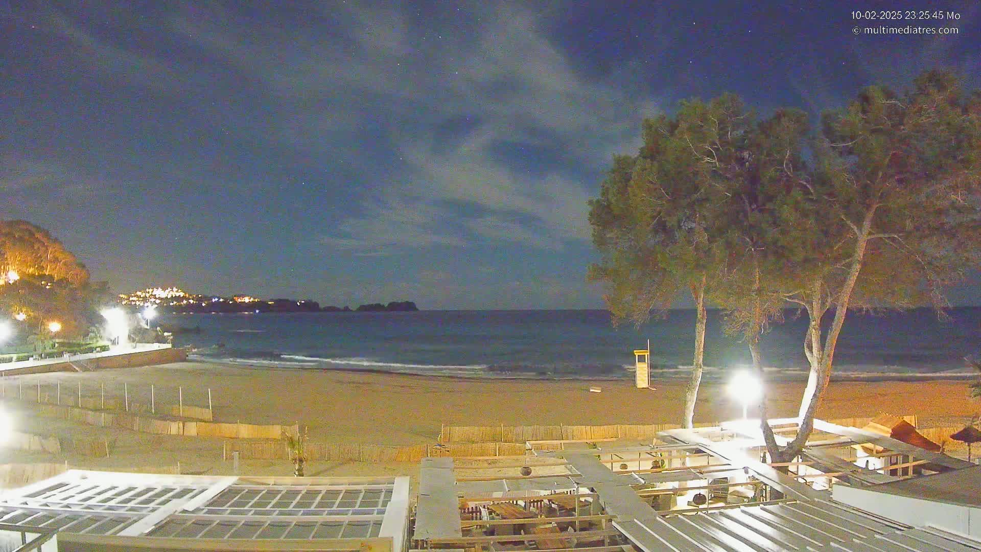 A nighttime beach scene under a partly cloudy sky shows a sandy beach, gentle waves, a lifeguard stand, several trees, and an outdoor restaurant with lights illuminating the area.