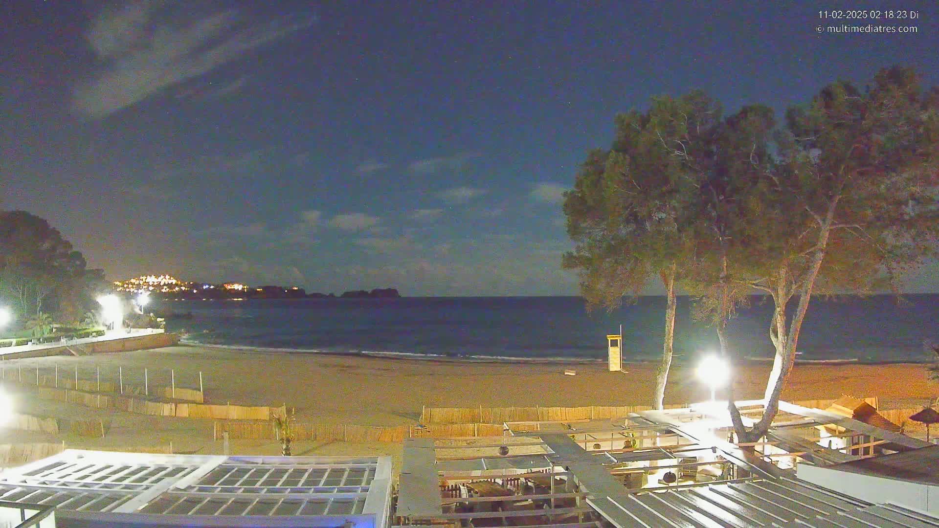 A nighttime view of a sandy beach with calm waves, illuminated by lights from a beachfront establishment, under a partly cloudy night sky.