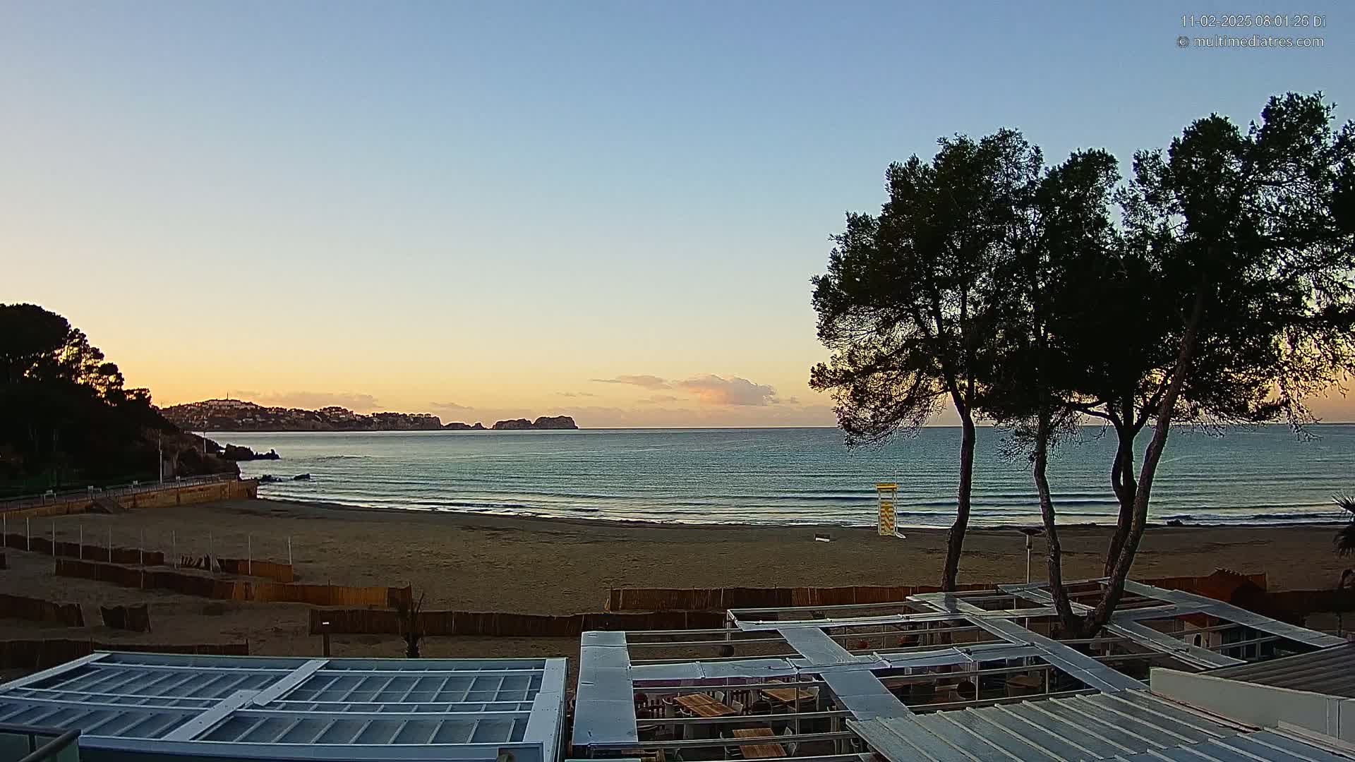 A sandy beach at sunrise, with calm ocean waves lapping the shore, overlooked by several pine trees and a partially constructed outdoor seating area.