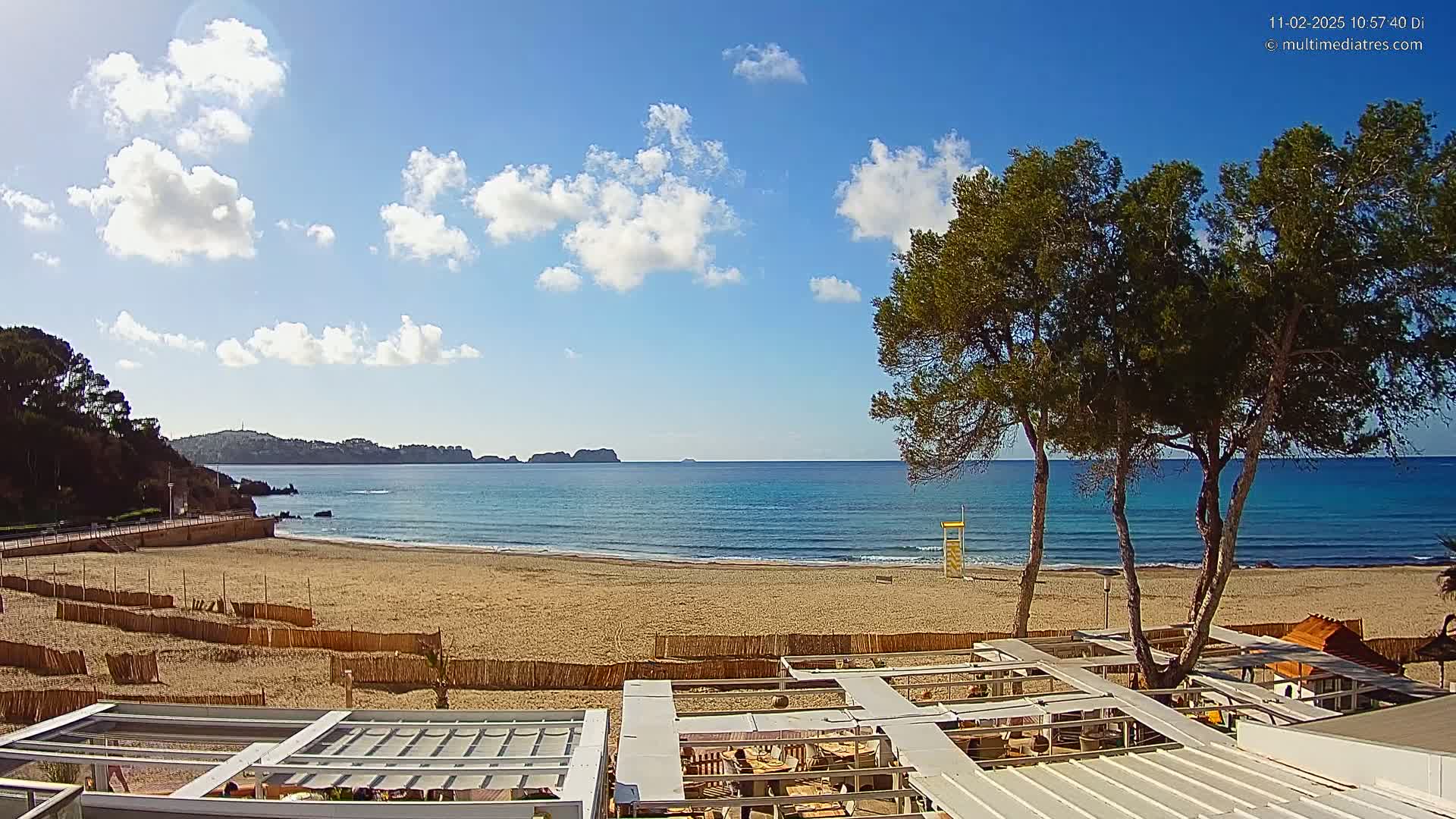 A sandy beach with calm, blue water stretches to a distant coastline under a mostly sunny sky with scattered clouds, and several pine trees stand in the foreground near an outdoor patio structure.