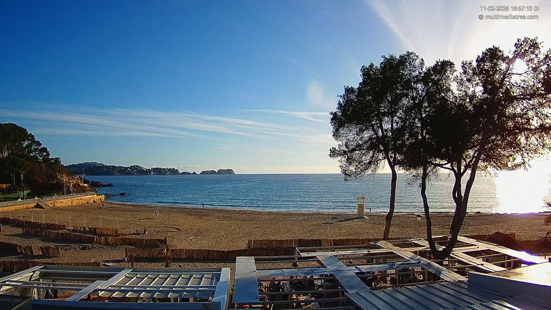 A sandy beach stretches along a calm, blue ocean under a clear, sunny sky, with several trees in the foreground and distant hills visible across the water.
