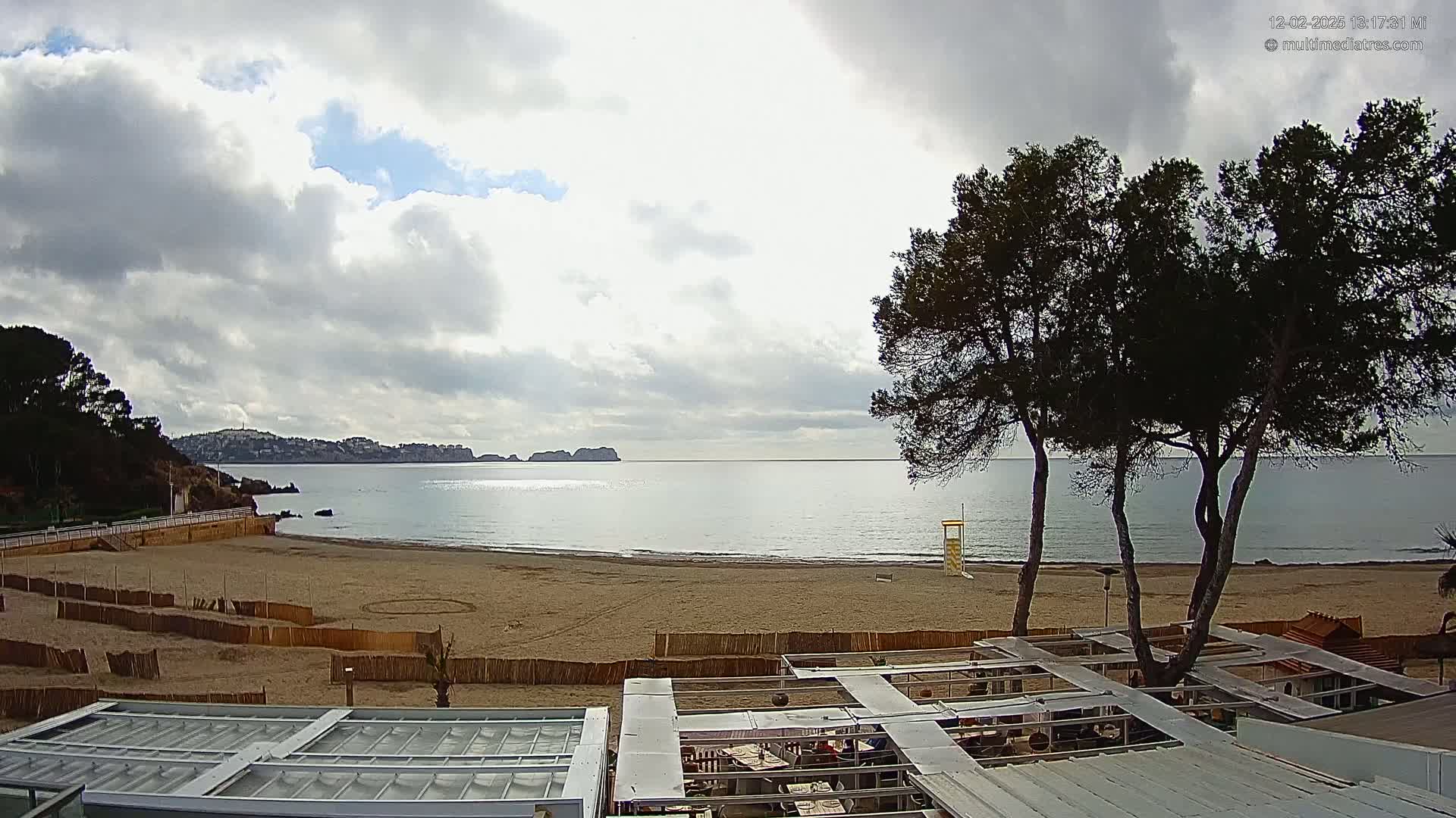 A sandy beach with calm water and a partly cloudy sky is viewed from above a beachfront restaurant, with several dark green trees in the foreground.