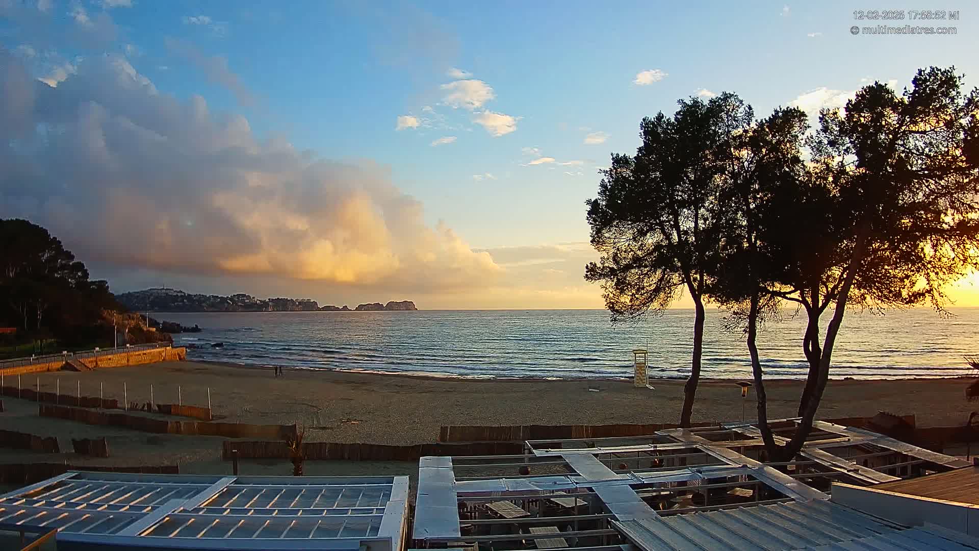 A sandy beach with calm waves at sunset under a partly cloudy sky, viewed from above a beachfront structure with several trees in the foreground.