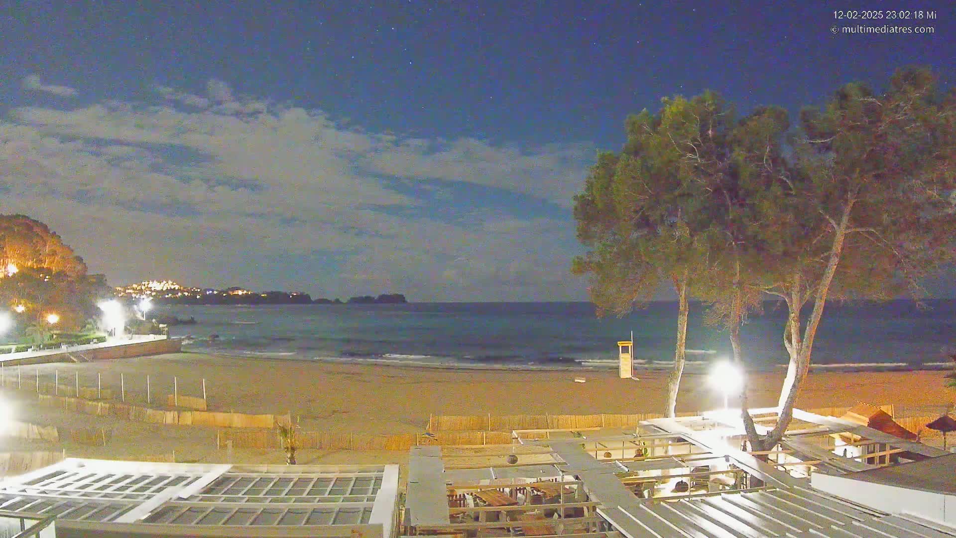 A nighttime view of a sandy beach with gentle waves, a lifeguard stand, and several illuminated trees overlooking a calm ocean under a partly cloudy sky.