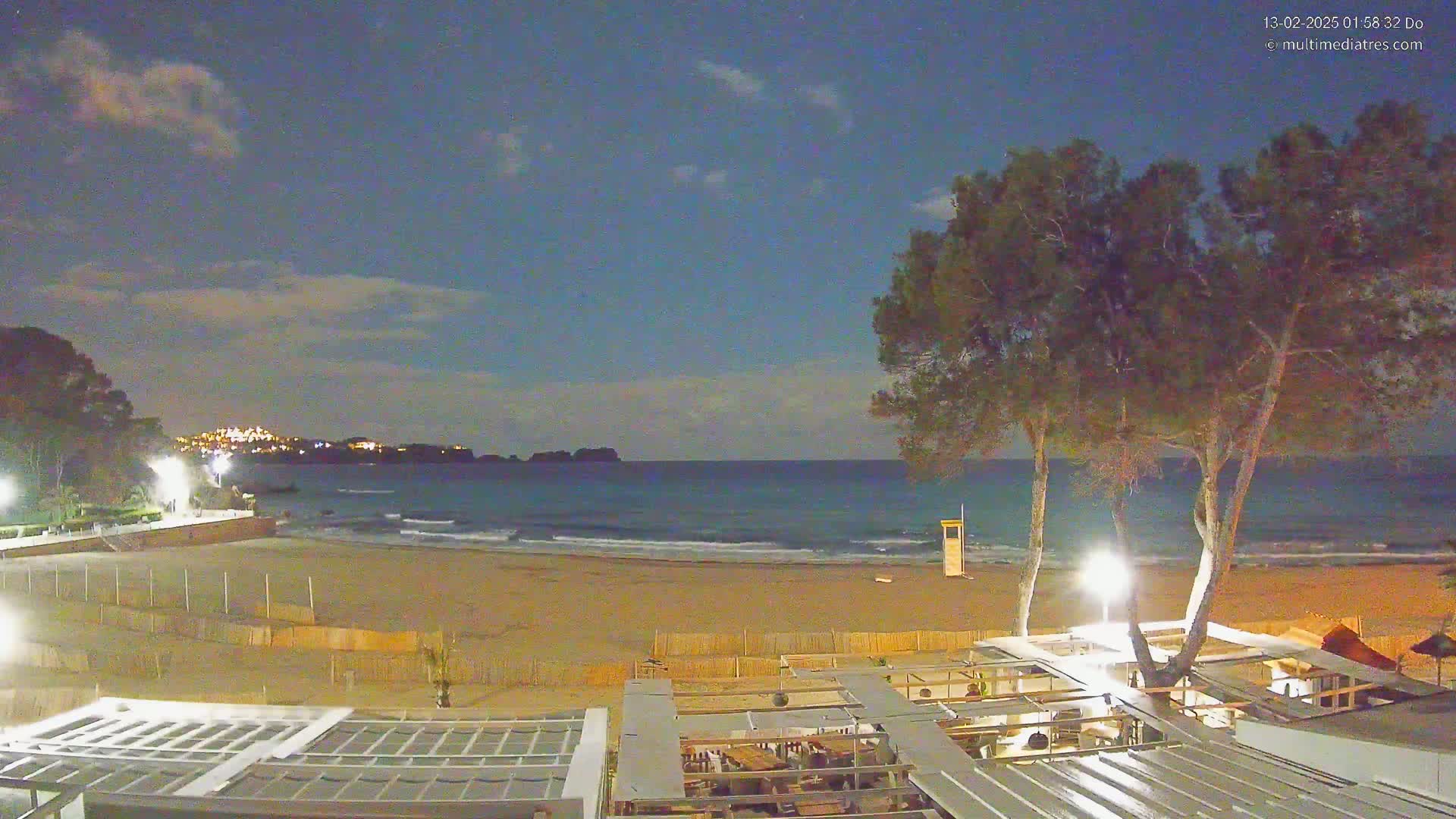A nighttime view of a sandy beach with gentle waves, illuminated by artificial lights, overlooking a calm sea under a partly cloudy sky.