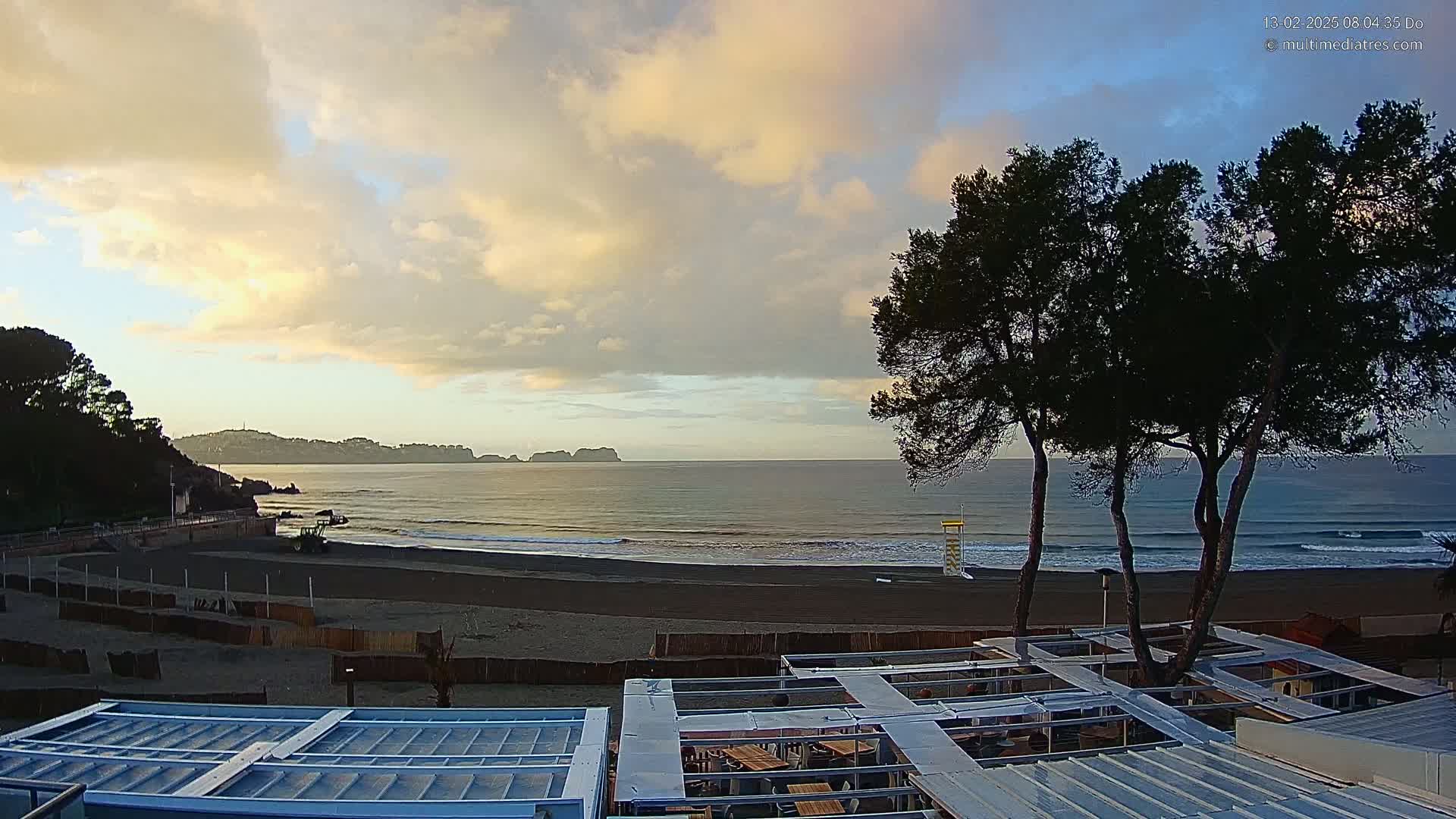 A sandy beach with gentle waves under a partly cloudy sunrise sky is viewed from above a partially covered outdoor patio area, with dark green trees in the foreground.