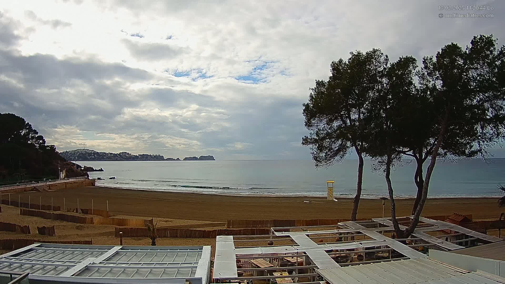 A sandy beach with gentle waves under a cloudy sky, viewed from a vantage point overlooking a beachfront structure with several pine trees in the foreground.