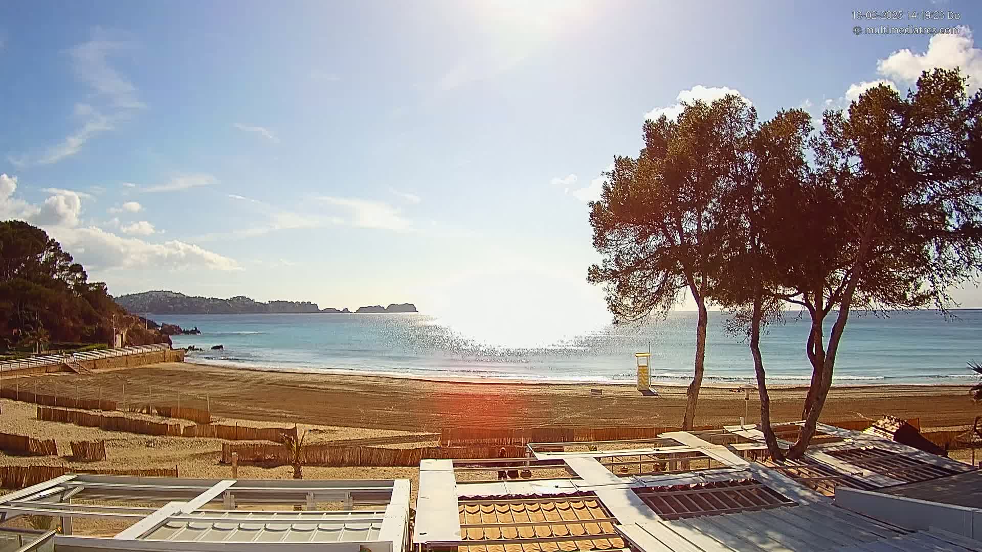 A sandy beach with calm, clear water under a mostly sunny sky is viewed from behind a row of buildings with retractable awnings; several pine trees stand between the buildings and the beach.