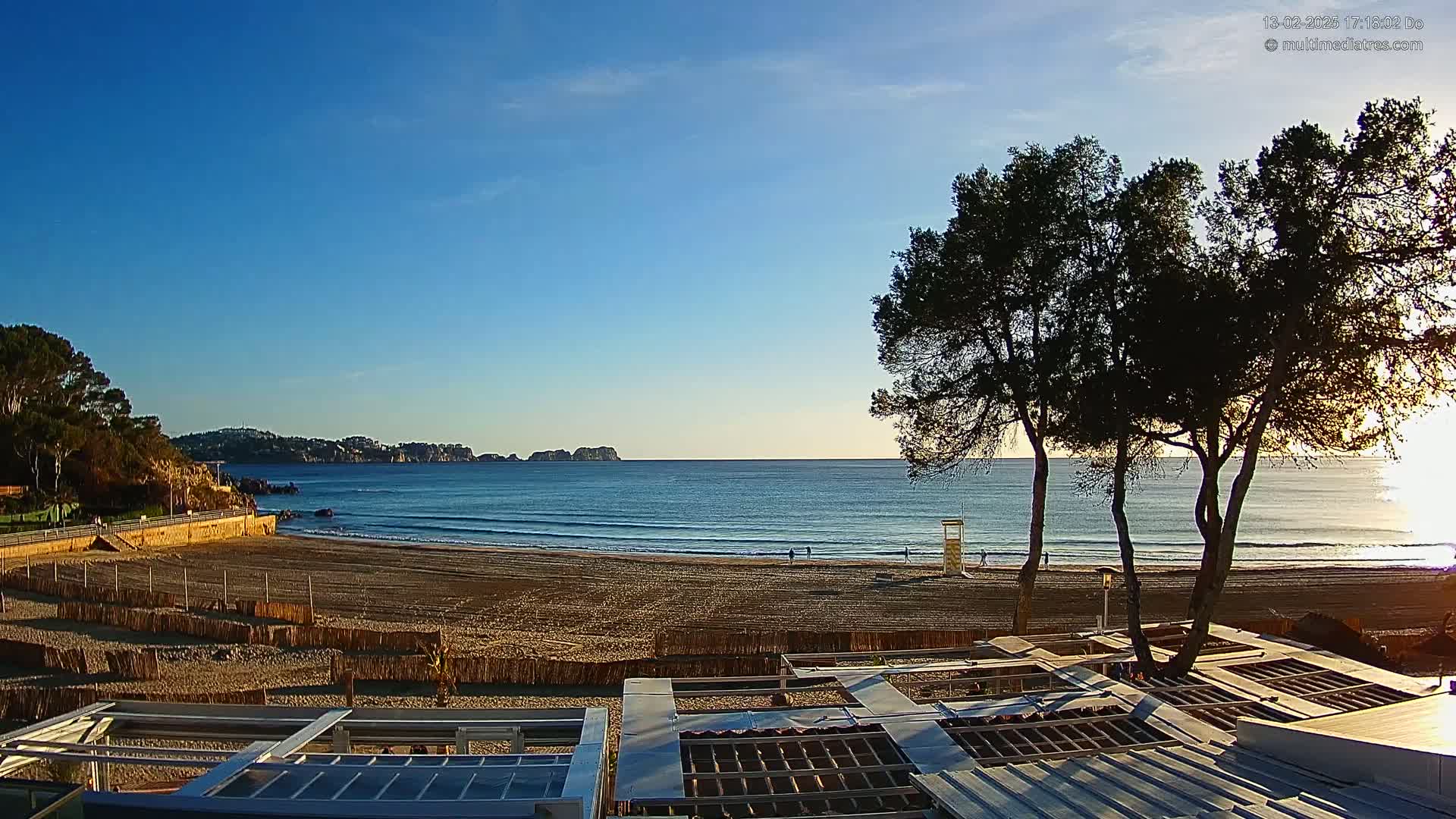 A sandy beach with calm waves under a clear blue sky is viewed from behind a structure with a corrugated metal roof, with a few people visible in the distance and dark trees in the foreground.