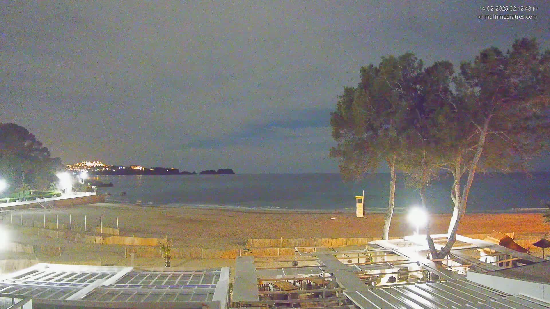 A nighttime view of a beach with calm water under a dusky sky, showing a sandy shore, beachfront structures, and tall trees.