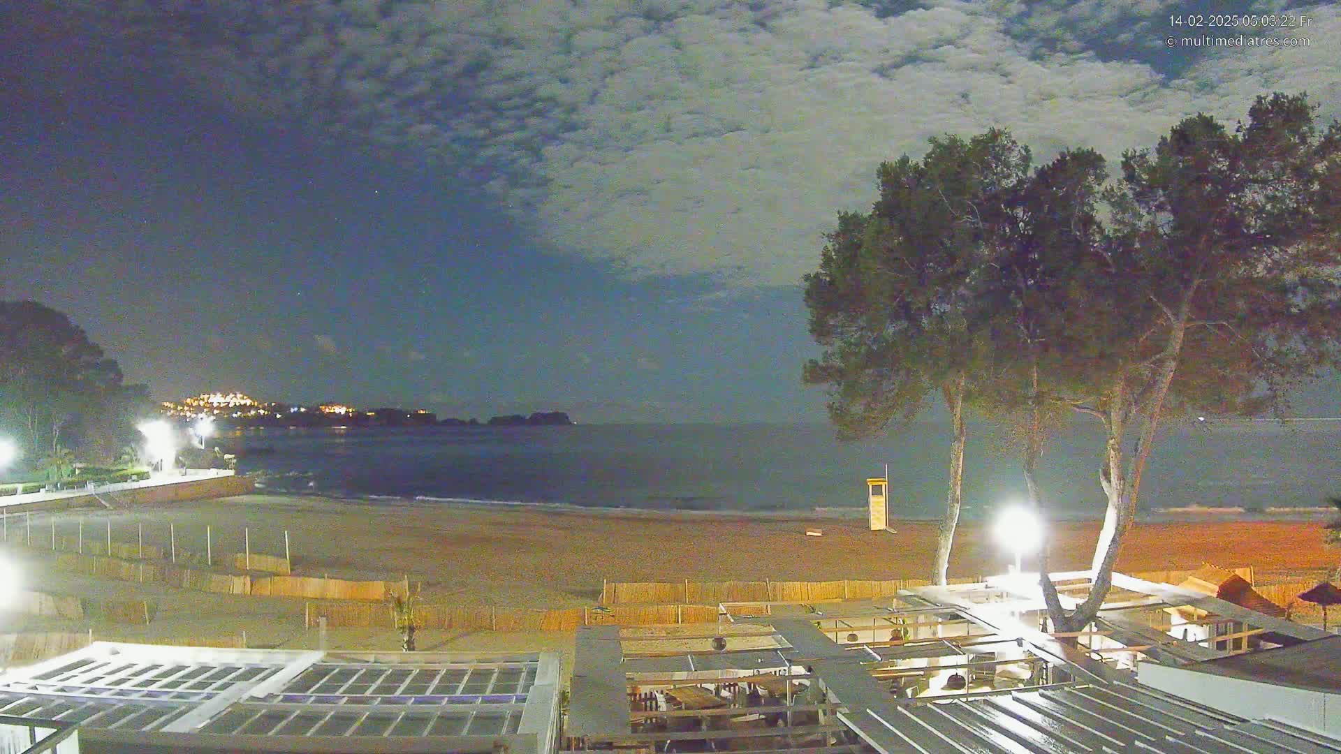 A nighttime view of a sandy beach with calm water, a few buildings, and trees under a partly cloudy sky.