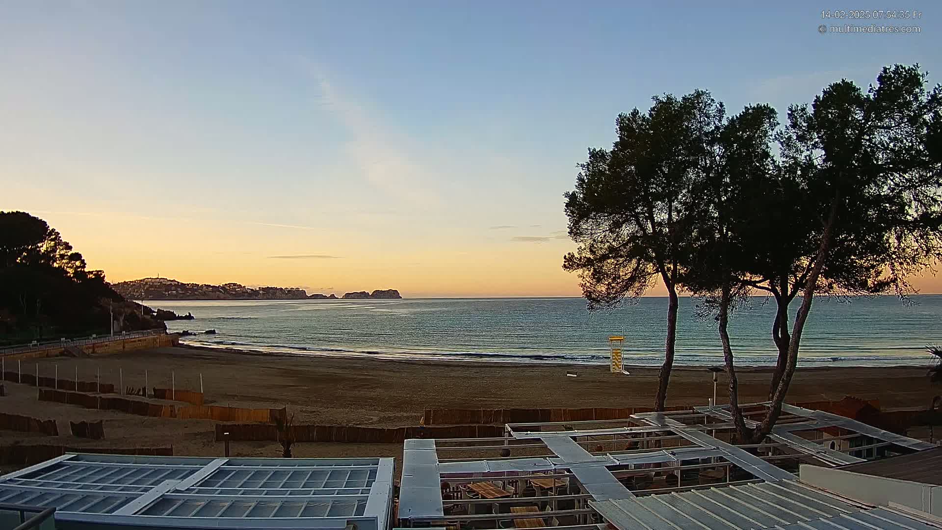 A sandy beach at sunrise, with calm ocean waves lapping the shore, is viewed from behind a beachfront structure, framed by several dark pine trees.