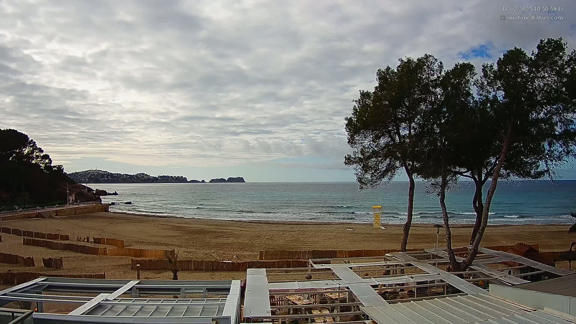 A sandy beach with gentle waves under a cloudy sky is viewed from above a partially covered outdoor seating area, with several pine trees in the foreground.