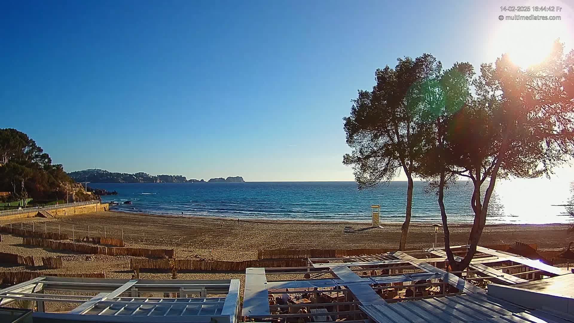 A sandy beach stretches towards a calm ocean under a clear, sunny sky, partially obscured by several pine trees and the roof of an outdoor structure.