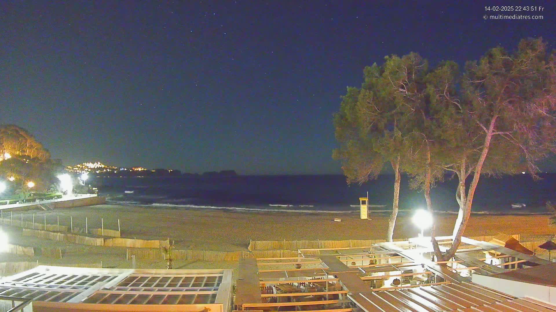 A night-time view of a beach with calm waves, illuminated by artificial lights, under a clear, starry sky.