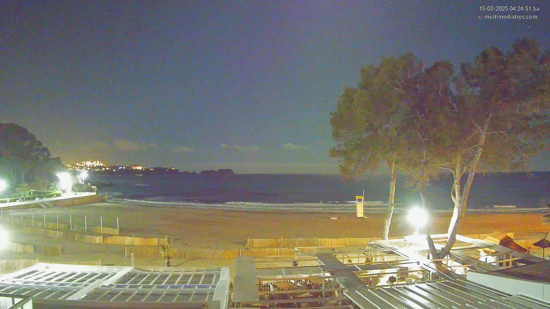 A nighttime view of a sandy beach with gentle waves, illuminated by artificial lights, under a partly cloudy sky with visible stars.