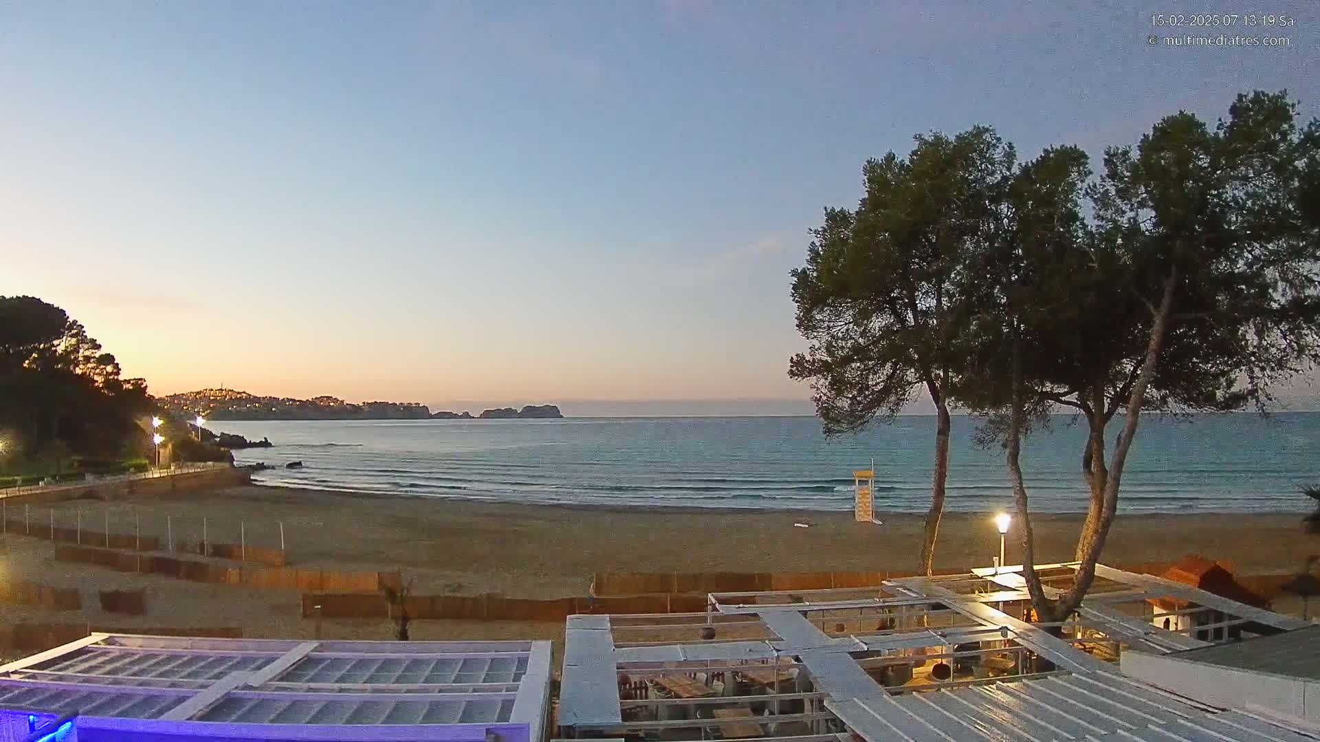 A sandy beach at dusk, with calm waves lapping the shore, is viewed from an elevated position overlooking a partially constructed beachfront structure and several pine trees.