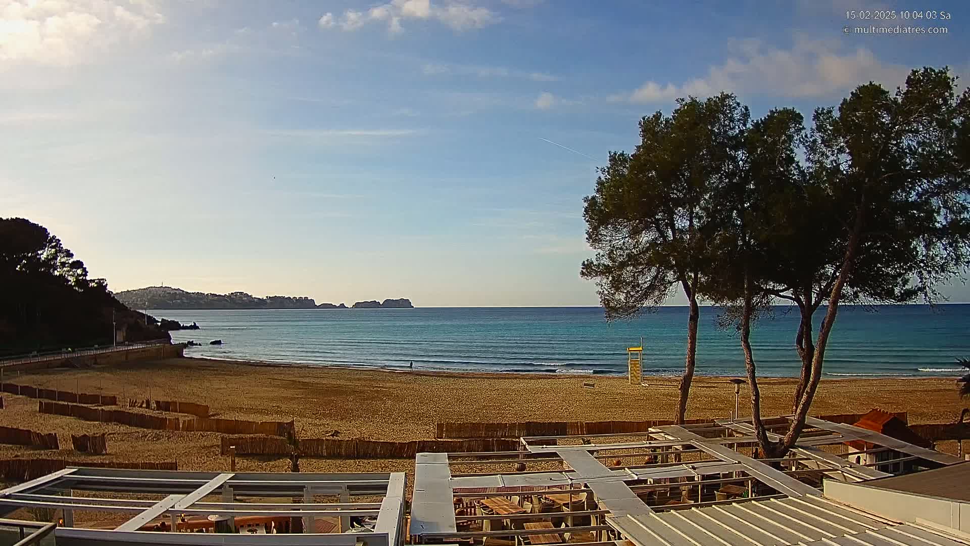 A sandy beach with gentle waves laps a calm turquoise sea under a mostly clear sky, with several pine trees in the foreground and a partially visible building.