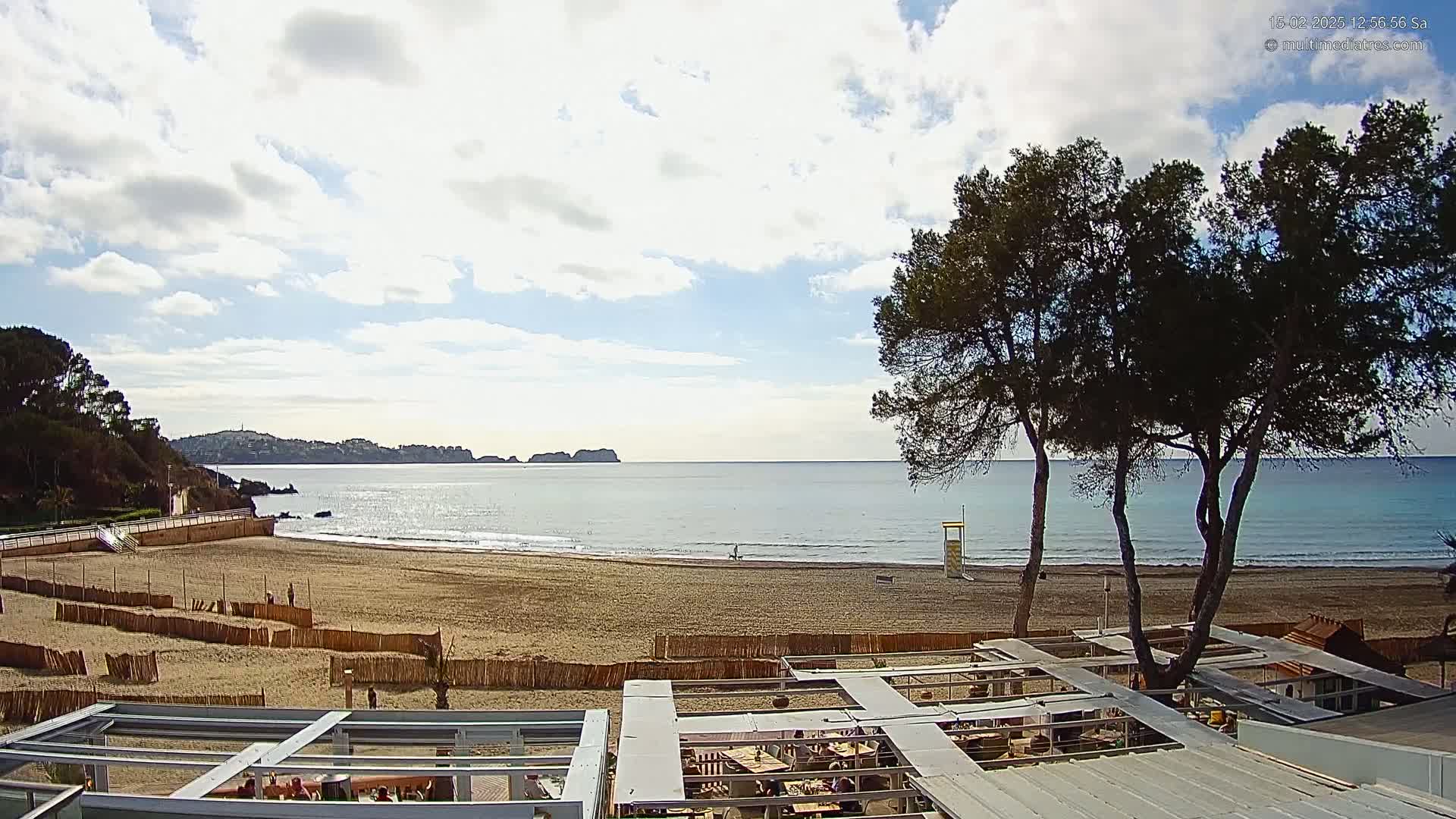 A sandy beach with calm ocean waters under a partly cloudy sky is viewed from a rooftop patio with several trees in the foreground.