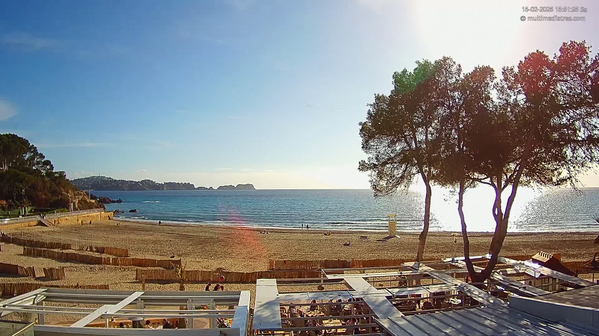 A sandy beach with calm, clear water and a few people is viewed from above, with several trees in the foreground under a mostly sunny sky.